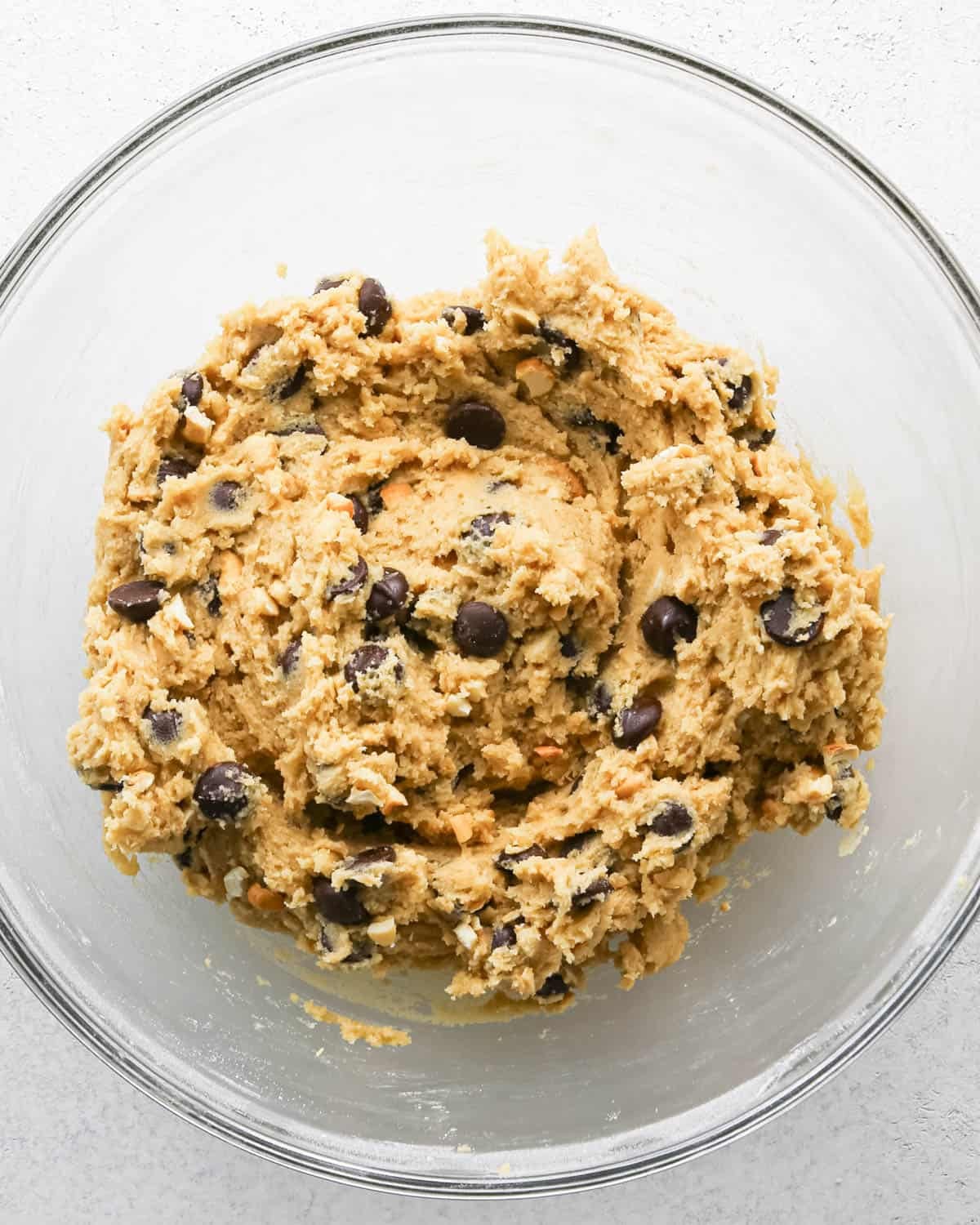 Making chocolate chip cashew cookies: overhead photo of chocolate chips and cashews added to the large clear mixing bowl with the cookie dough and mixed together.