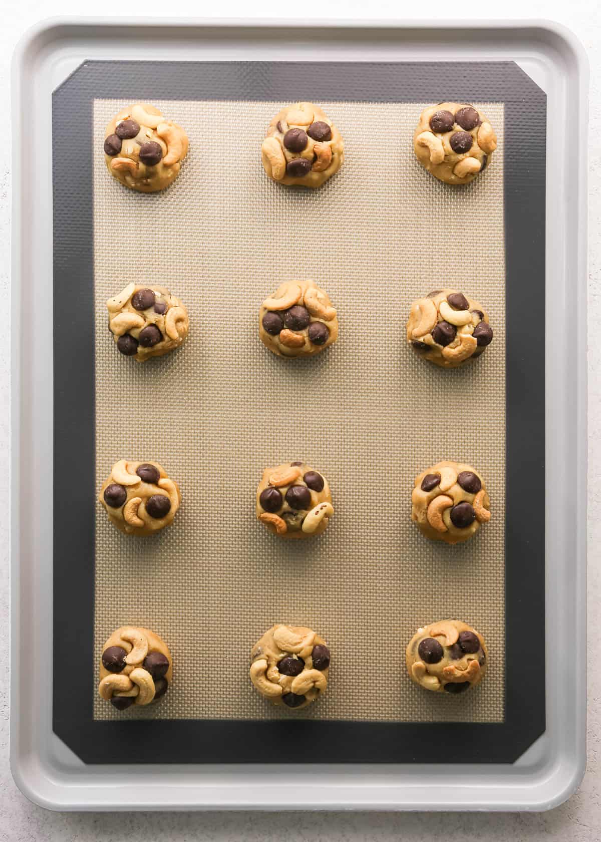 Making chocolate chip cashew cookies: overhead photo of 12 balls of cookie dough scooped, rolled, and placed onto a baking sheet on a countertop. Whole cashews have been added to the top of each cookie.