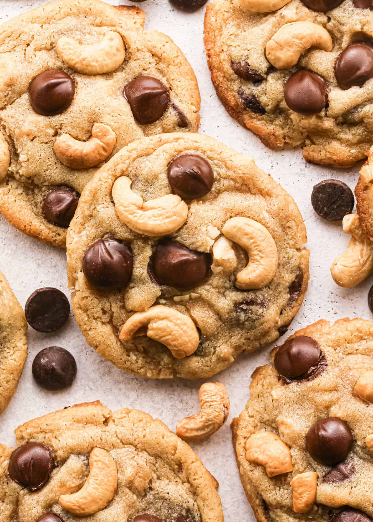 Overhead photo of golden-brown chocolate chip cashew cookies on parchment paper, studded with large chocolate chips and whole roasted cashews, with a few loose chocolate chips and cashews scattered around the cookies.