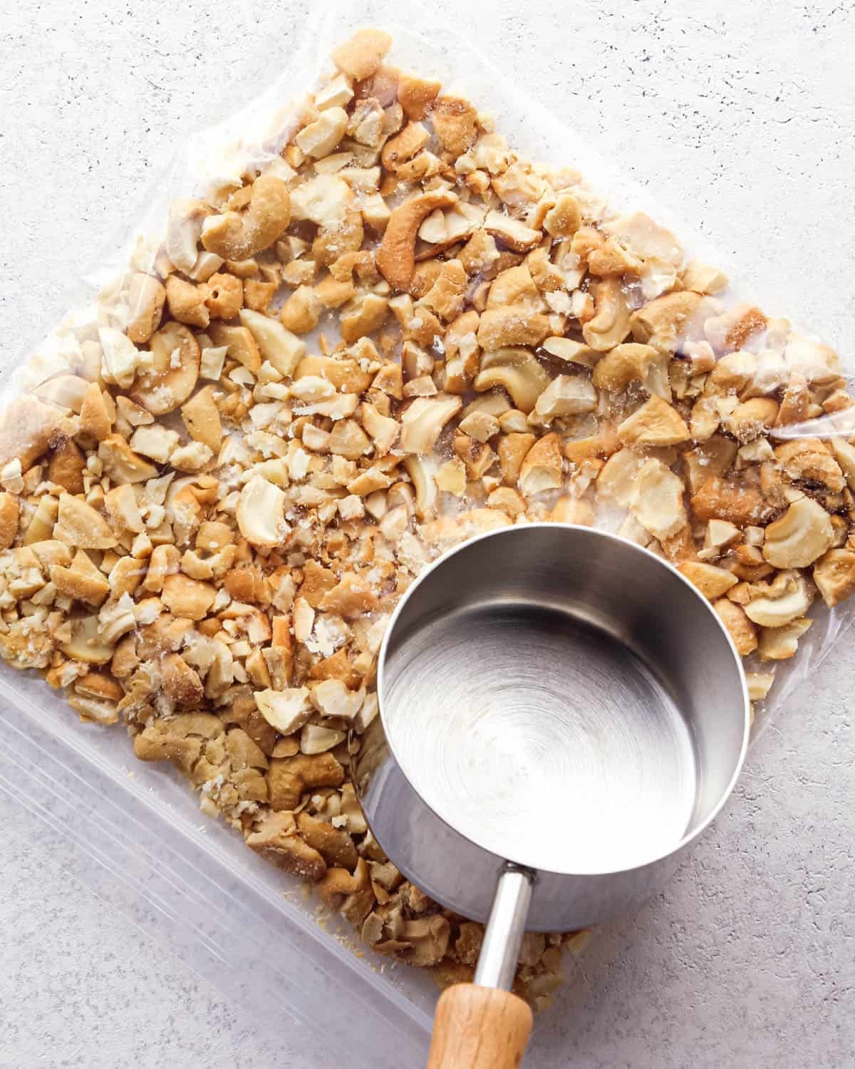 Making chocolate chip cashew cookies: overhead photo of a gallon plastic bag filled with cashews and being crushed inside the bag with a metal measuring cup.