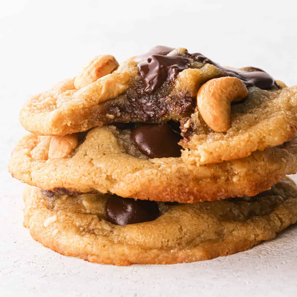 Close-up photo of a stack of 3 golden-brown chocolate chip cashew cookies on a white surface, studded with large chocolate chips and whole roasted cashews.