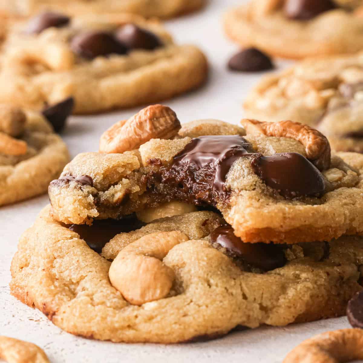 Up-close photo of a surface full of chocolate chip cashew cookies. A bite is taken out of one of the cookies to show the melty chocolate, the crispy outside, and the chewy, perfectly gooey inside of the cookie. Extra chocolate chips and cashews surround the cookies on a white countertop.