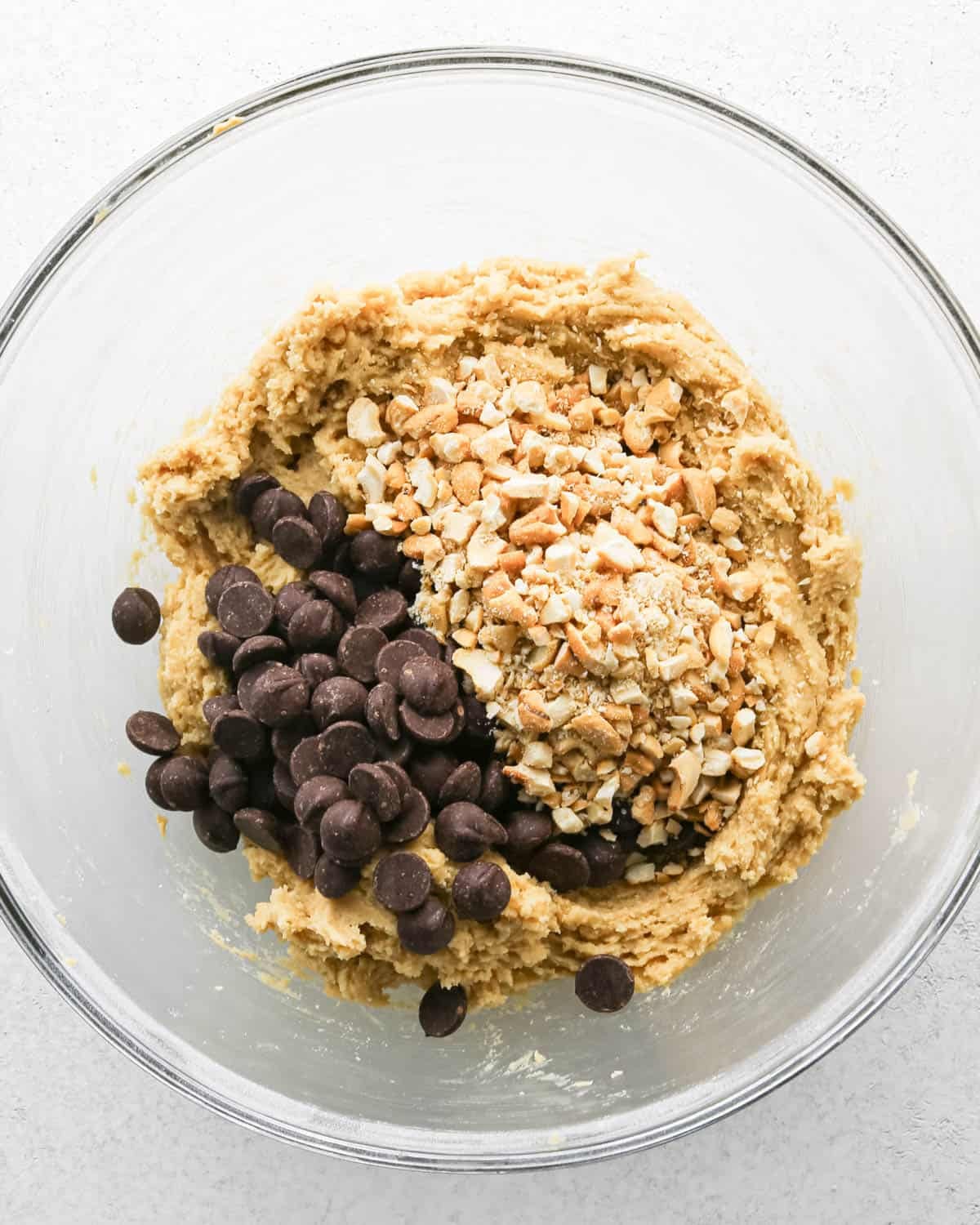 Making chocolate chip cashew cookies: overhead photo of chocolate chips and cashews added to the large clear mixing bowl with the cookie dough.