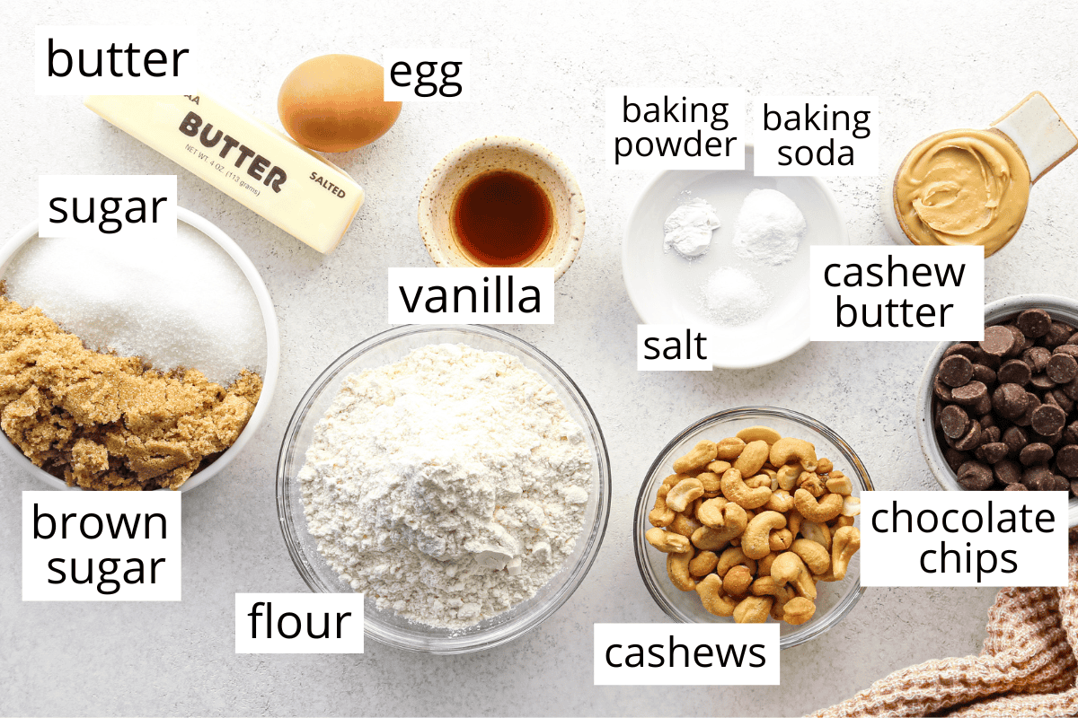 Overhead view of ingredients for chocolate chip cashew cookies arranged on a light countertop, including butter, egg, sugar, brown sugar, vanilla, flour, baking powder, baking soda, salt, cashew butter, cashews, and chocolate chips.