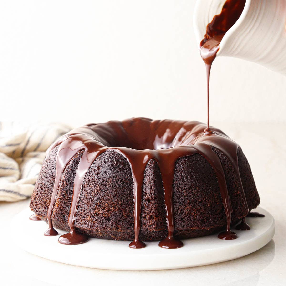 chocolate glaze being poured from a white container onto a chocolate bundt cake. the glaze is dripping down the sides onto a round white marble serving plate