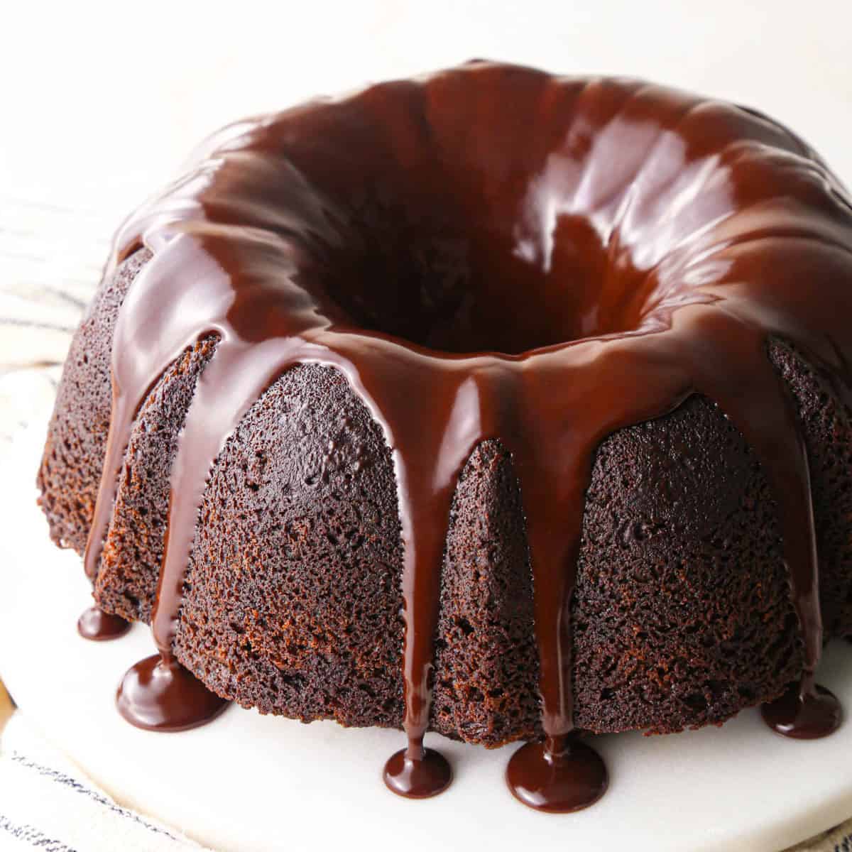 a chocolate bundt cake on a white cake plate topped with chocolate glaze