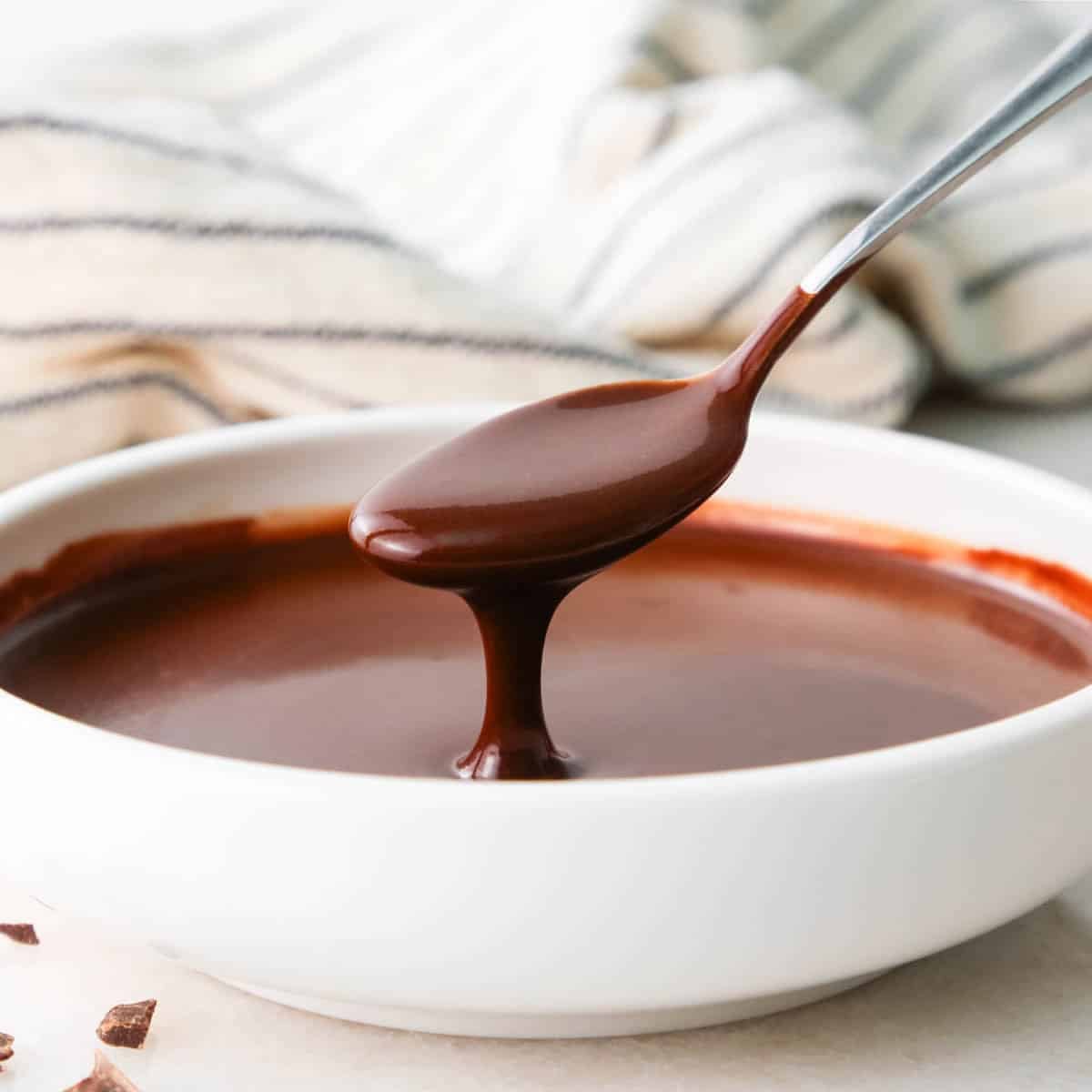 chocolate glaze in a white bowl with a spoon taking a scoop out of it and some of the glaze dripping back into the bowl