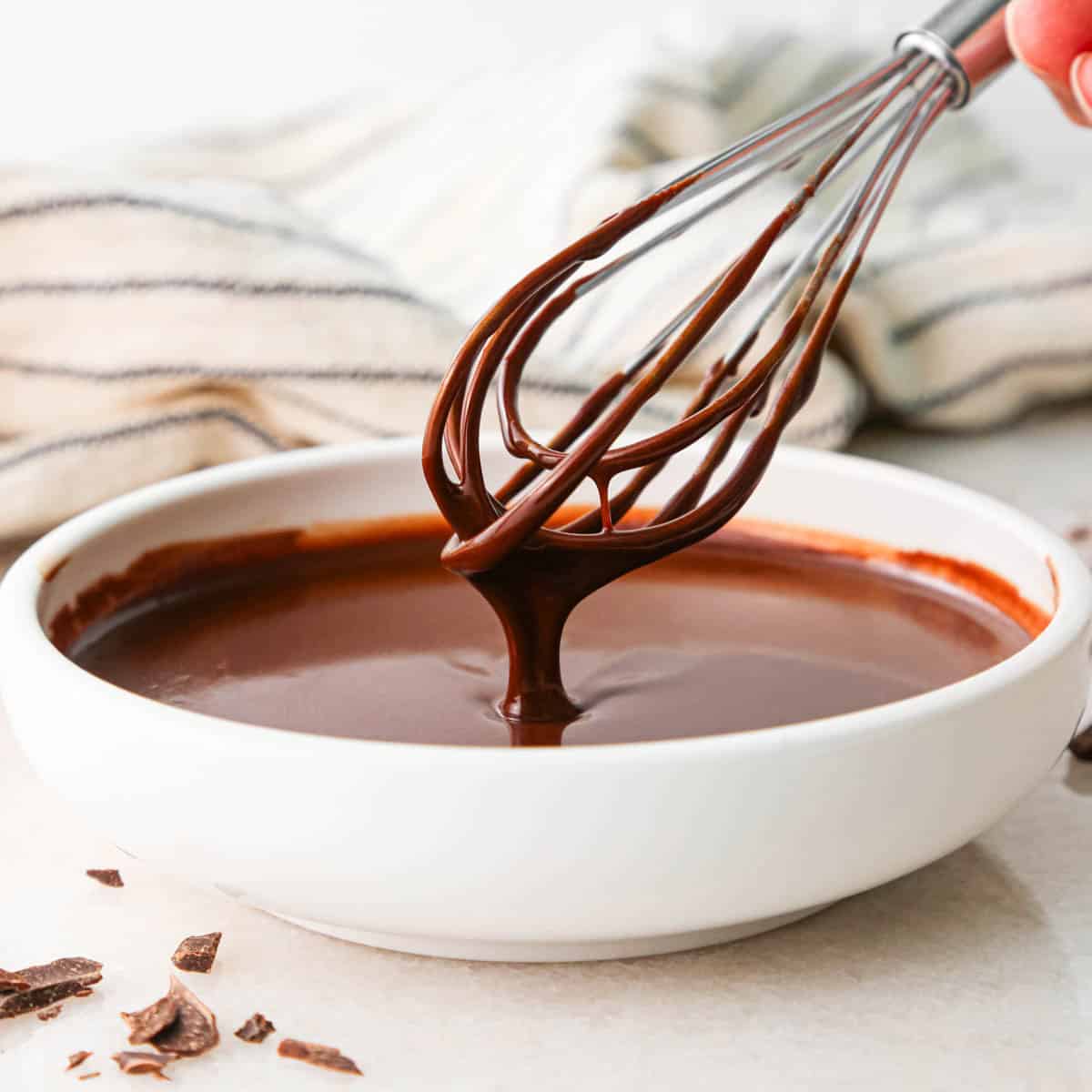 chocolate glaze in a white bowl with a wire whisk covered in glaze with some dripping off of it back into the bowl