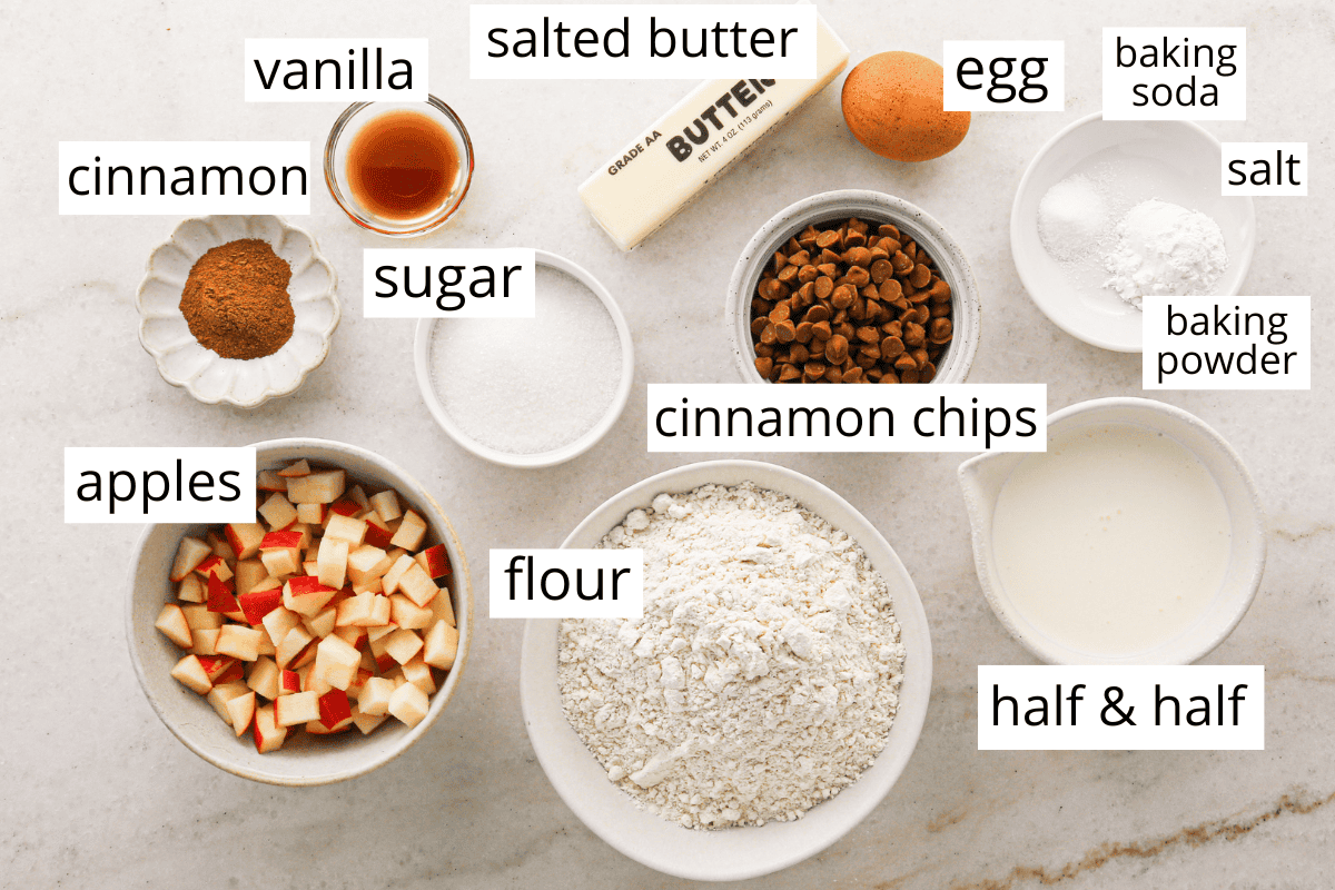 overhead photo of the ingredients in this cinnamon scones recipe all in bowls and labeled like flour, apples, cinnamon chips, and more.