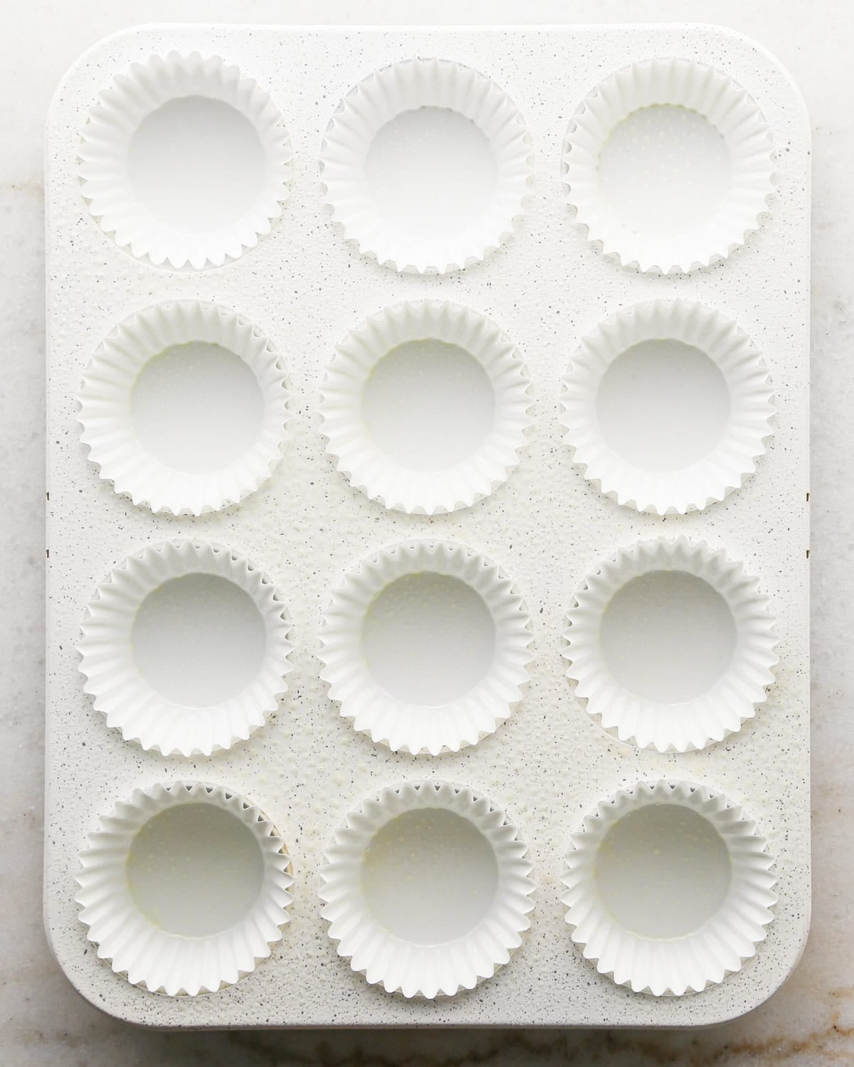 Making coconut cupcakes: overhead photo of a white speckled 12-cup muffin tin lined with white paper liners and sitting on a countertop.
