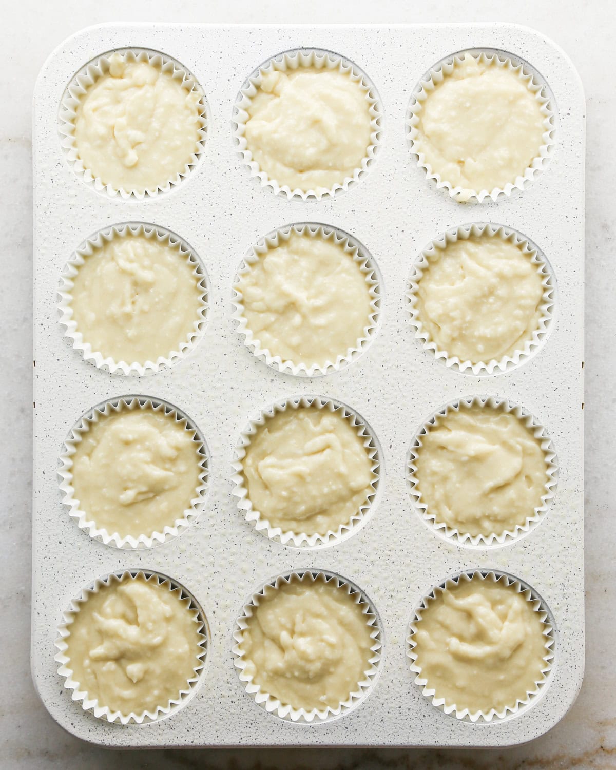 Making coconut cupcakes: overhead photo of a white speckled 12-cup muffin tin lined with white paper liners, each of which has been filled with cupcake batter.