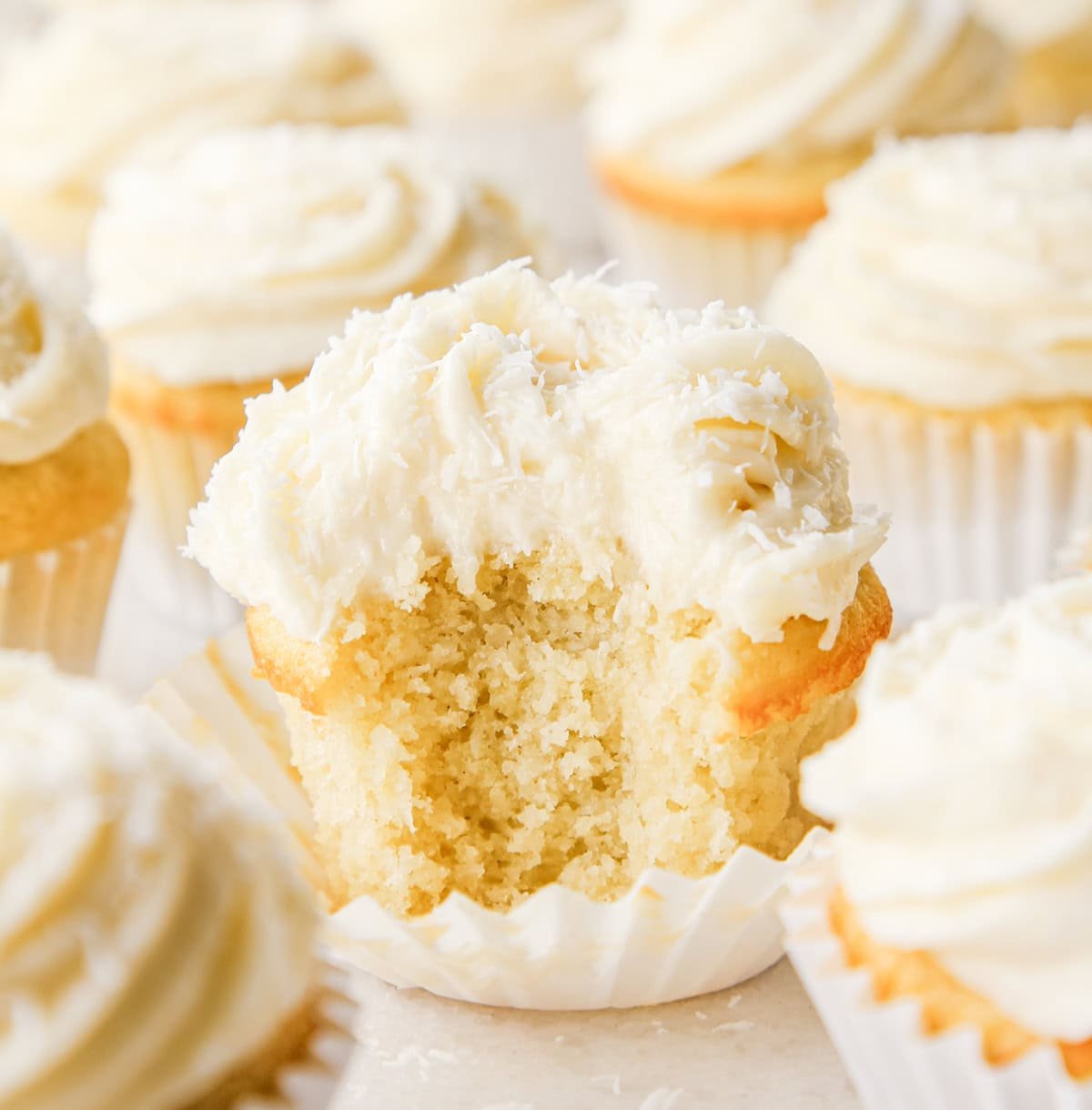 Close-up of a coconut cupcake with a bite taken out, showing a moist vanilla crumb topped with creamy coconut frosting and shredded coconut, in a white paper liner. Other cupcakes surround it on a light surface.