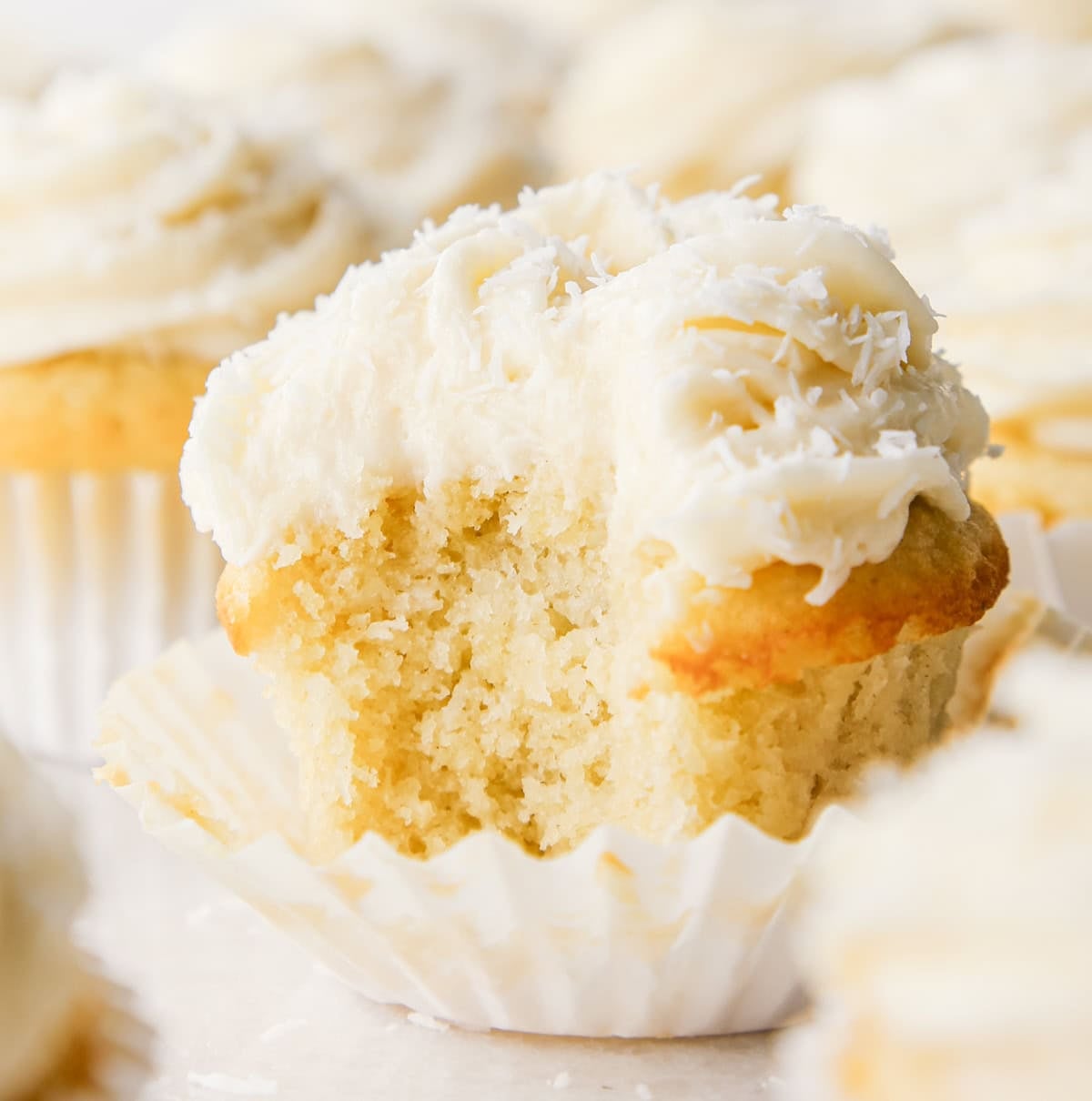 Close-up of a coconut cupcake with a bite taken out, showing a moist vanilla crumb topped with creamy coconut frosting and shredded coconut, in a white paper liner. Other cupcakes surround it on a light surface.