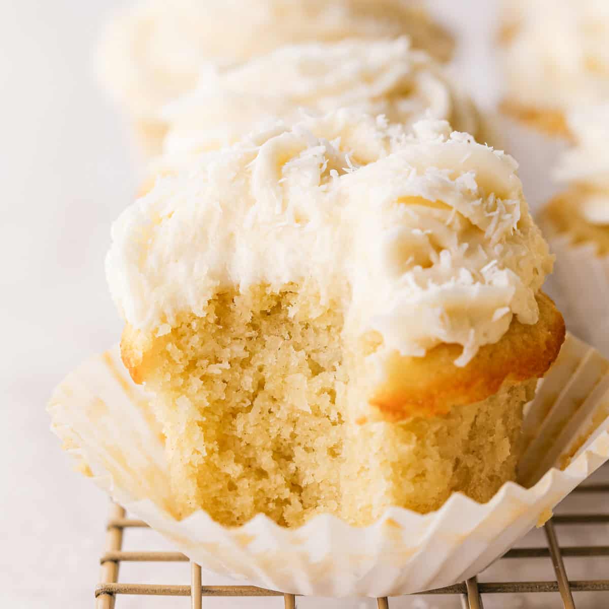 Close-up of a coconut cupcake with a bite taken out, showing a moist vanilla crumb topped with creamy coconut frosting and shredded coconut, in a white paper liner and sitting on a light-colored wire cooling rack.