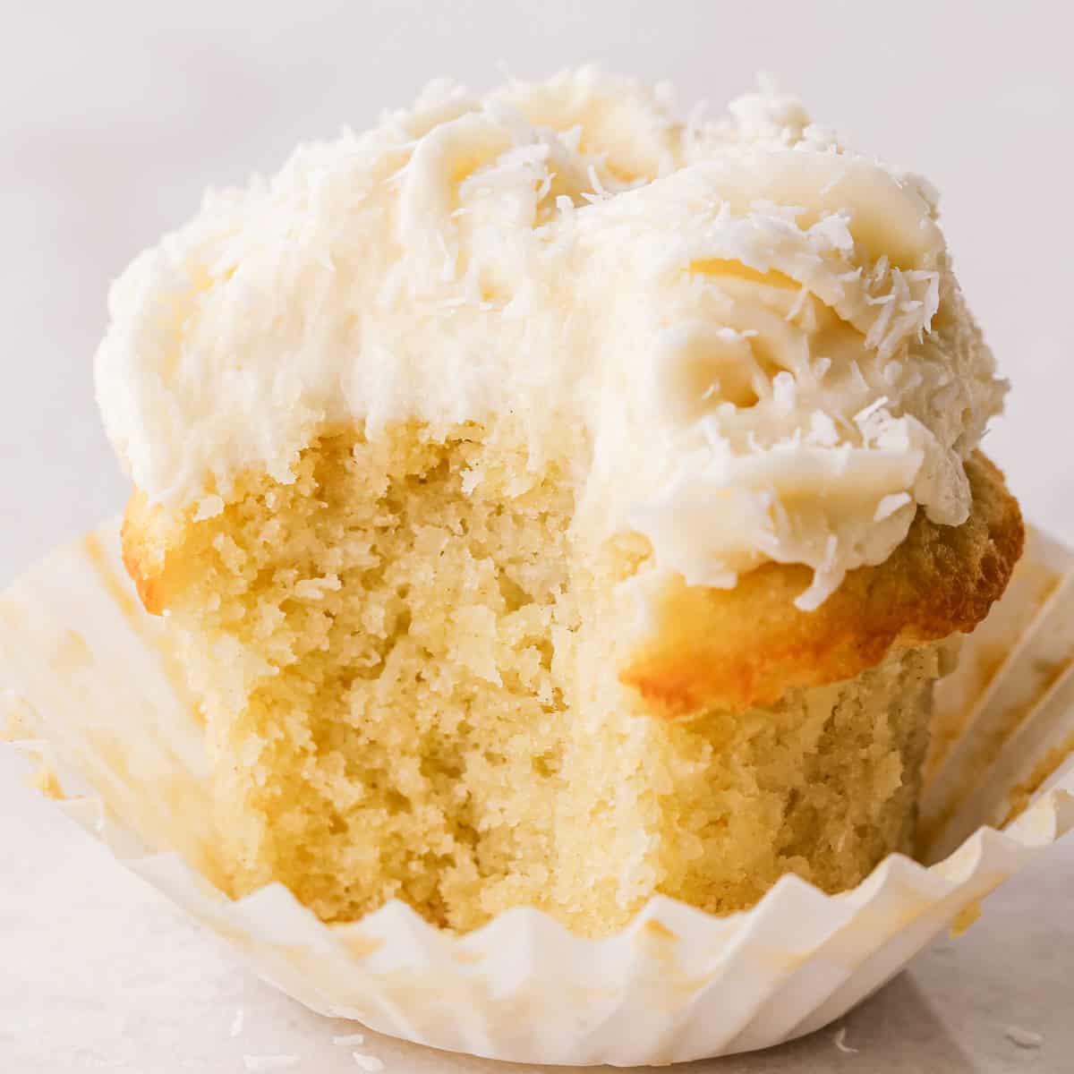 Close-up of a coconut cupcake with a bite taken out, showing a moist vanilla crumb topped with creamy coconut frosting and shredded coconut, in a white paper liner.