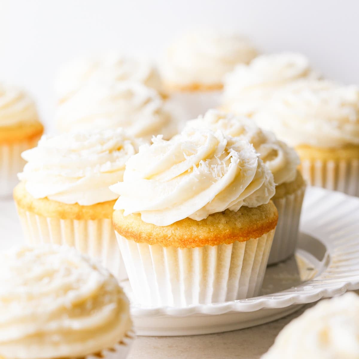 Close-up of a coconut cupcake, showing a moist vanilla crumb topped with creamy coconut frosting and shredded coconut, in a white paper liner. Other cupcakes surround it on a white plate.