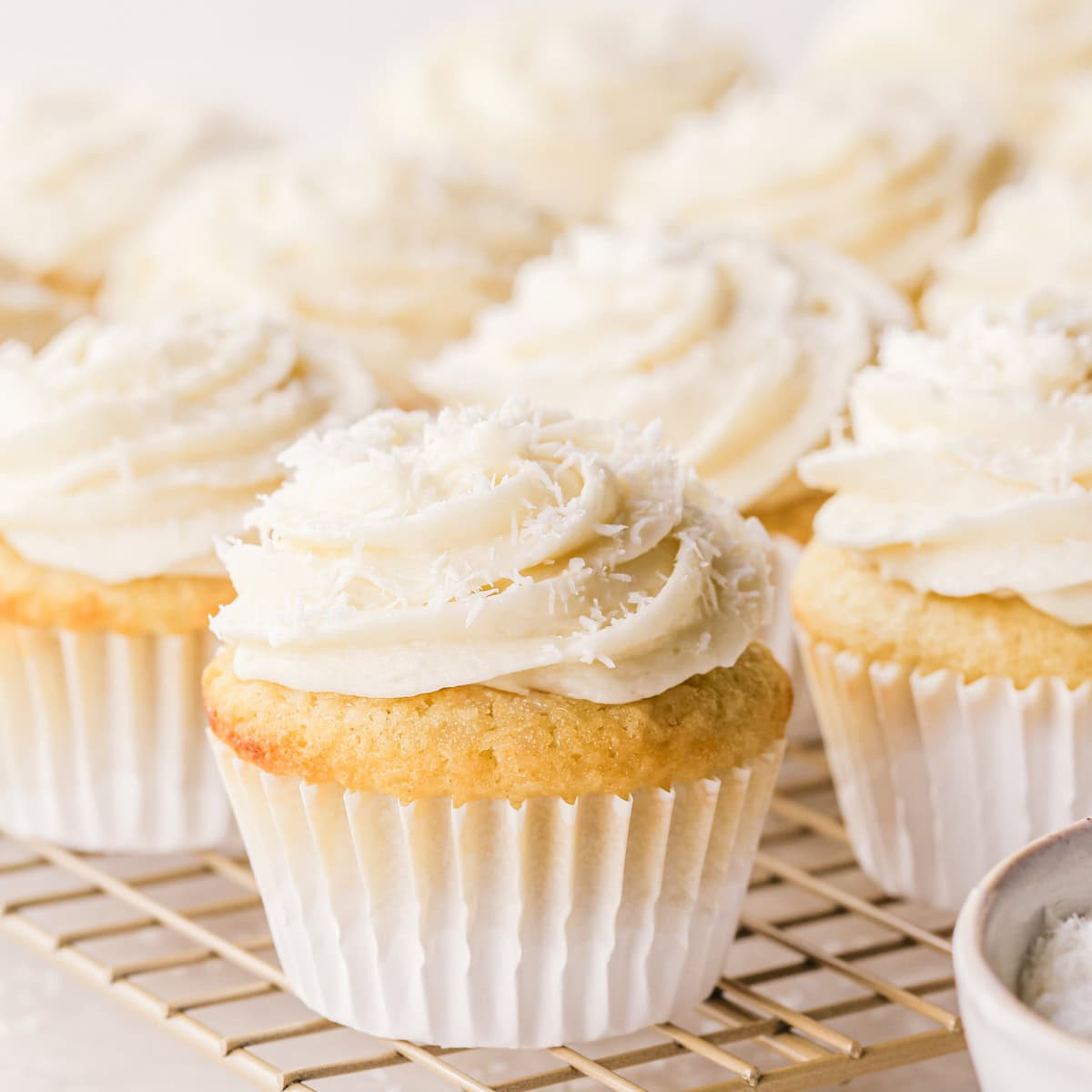 Close-up of a coconut cupcake, showing a moist vanilla crumb topped with creamy coconut frosting and shredded coconut, in a white paper liner. Other cupcakes surround it on a light-colored wire cooling rack.