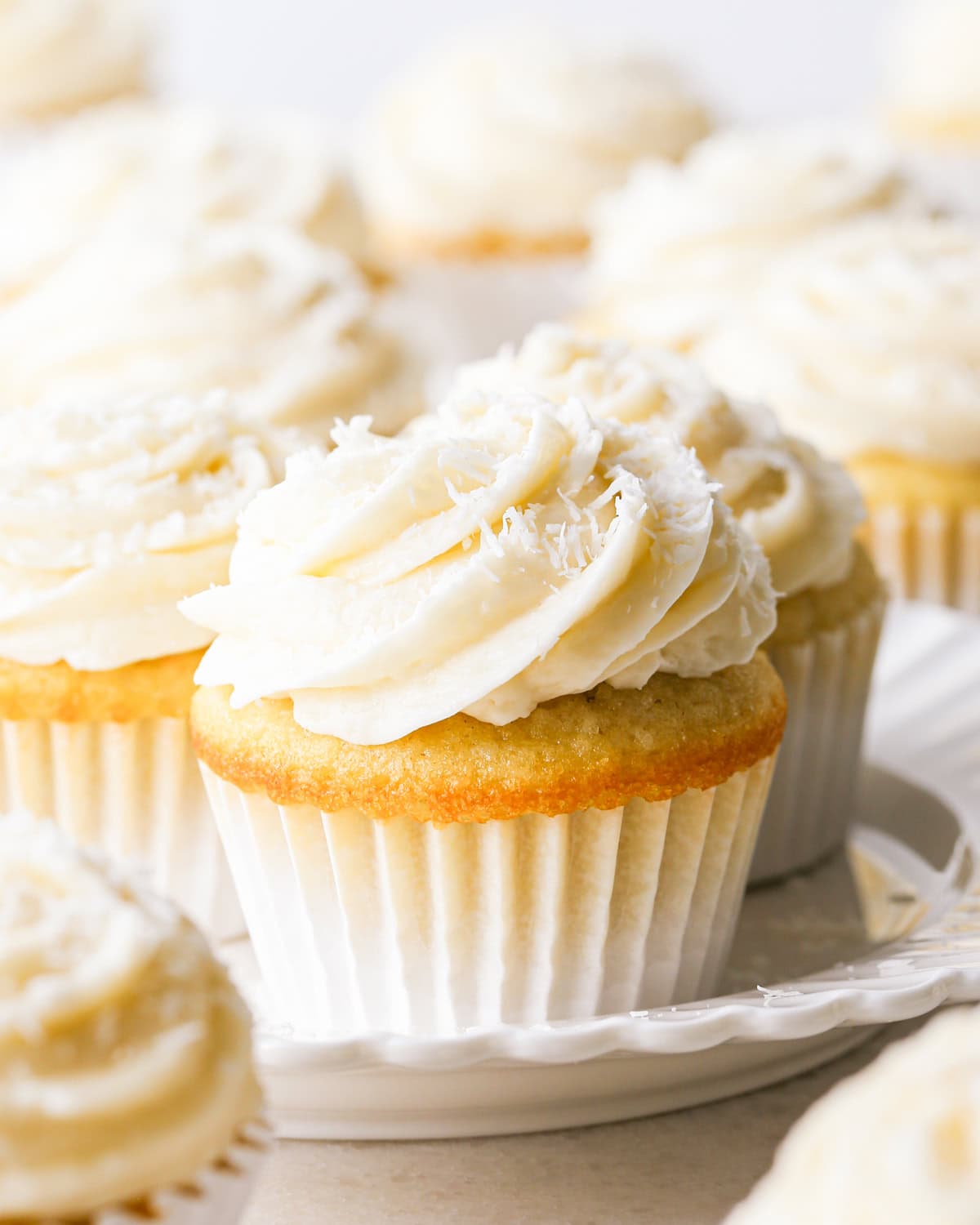 Close-up of a coconut cupcake, showing a moist vanilla crumb topped with creamy coconut frosting and shredded coconut, in a white paper liner. Other cupcakes surround it on a white plate.