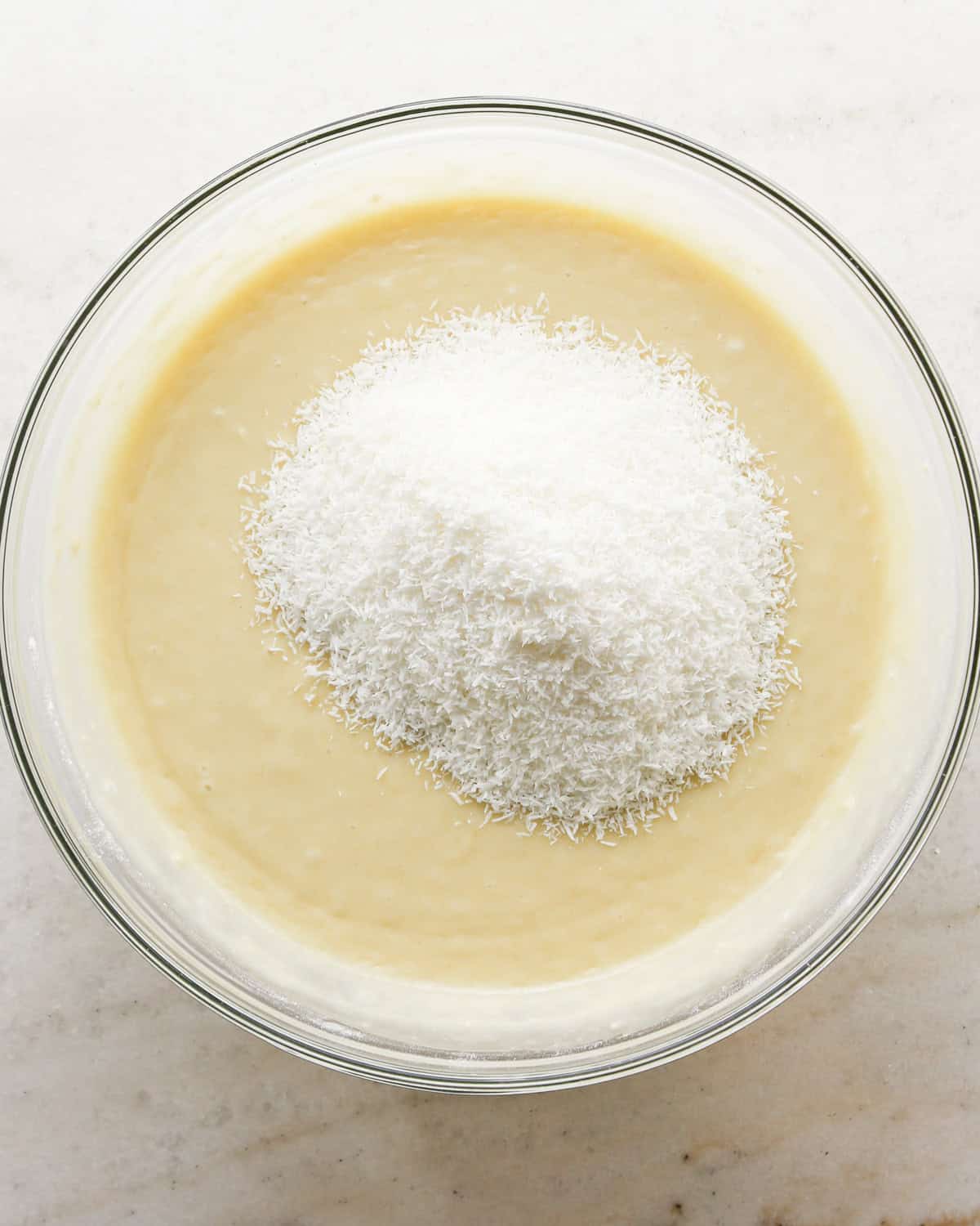 Making coconut cupcakes: overhead photo of shredded coconut being added to the cupcake batter in a clear mixing bowl.
