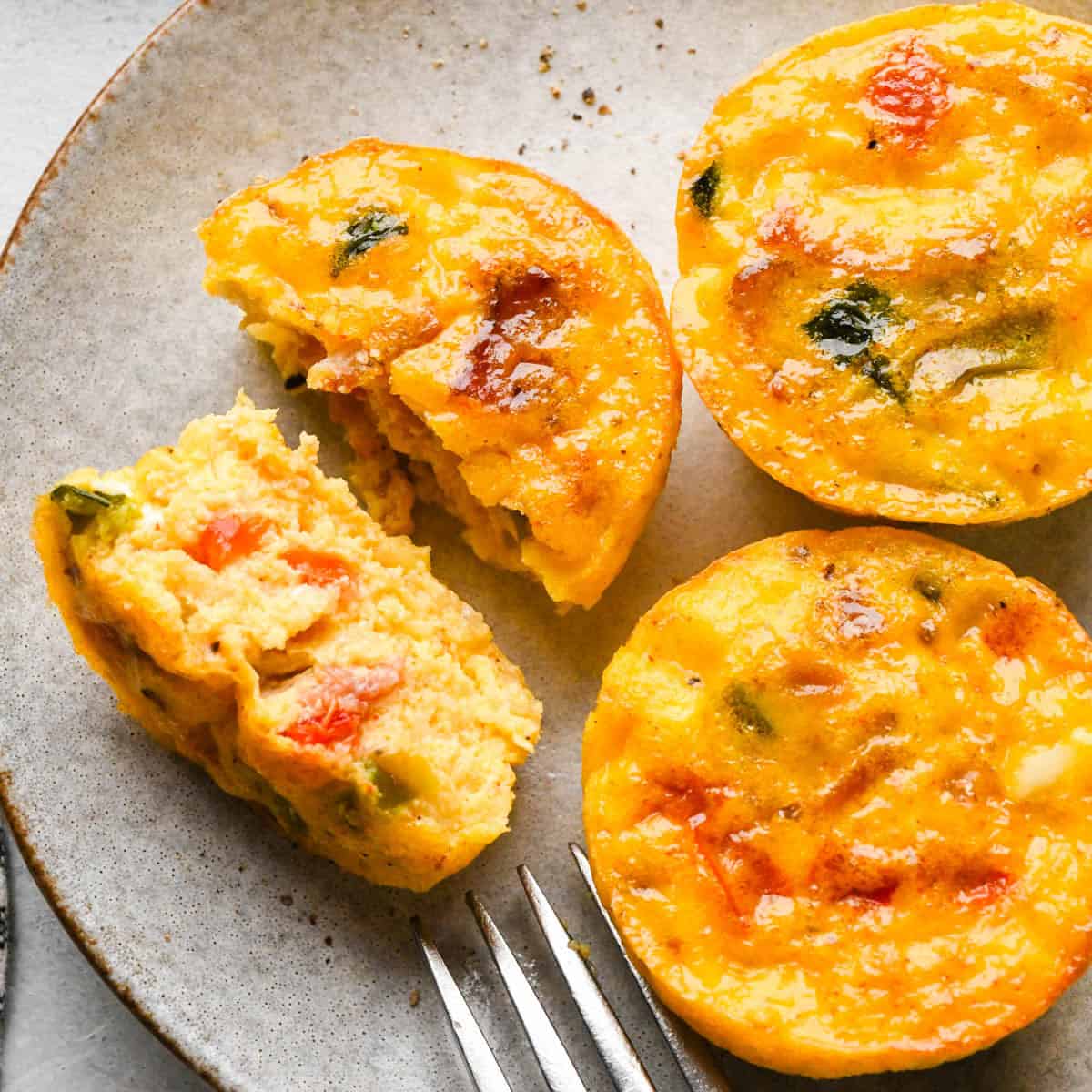 3 egg bites with cottage cheese on a gray pottery plate, one is cut in half and one of the halves is on its' side so the fluffy texture and vegetables inside are visible. There's also a fork on the plate