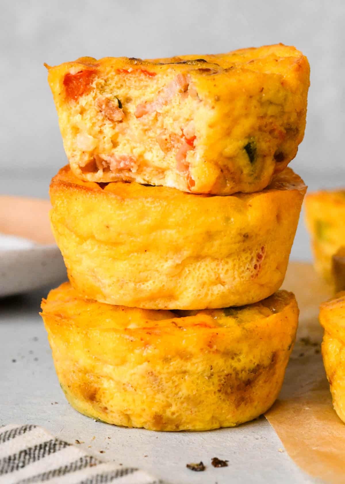 a stack of 3 cottage cheese egg bites, the top one has a bite taken out of it, surrounded by two other egg bites with cottage cheese on parchment paper and a bowl of salt and pepper in the background.