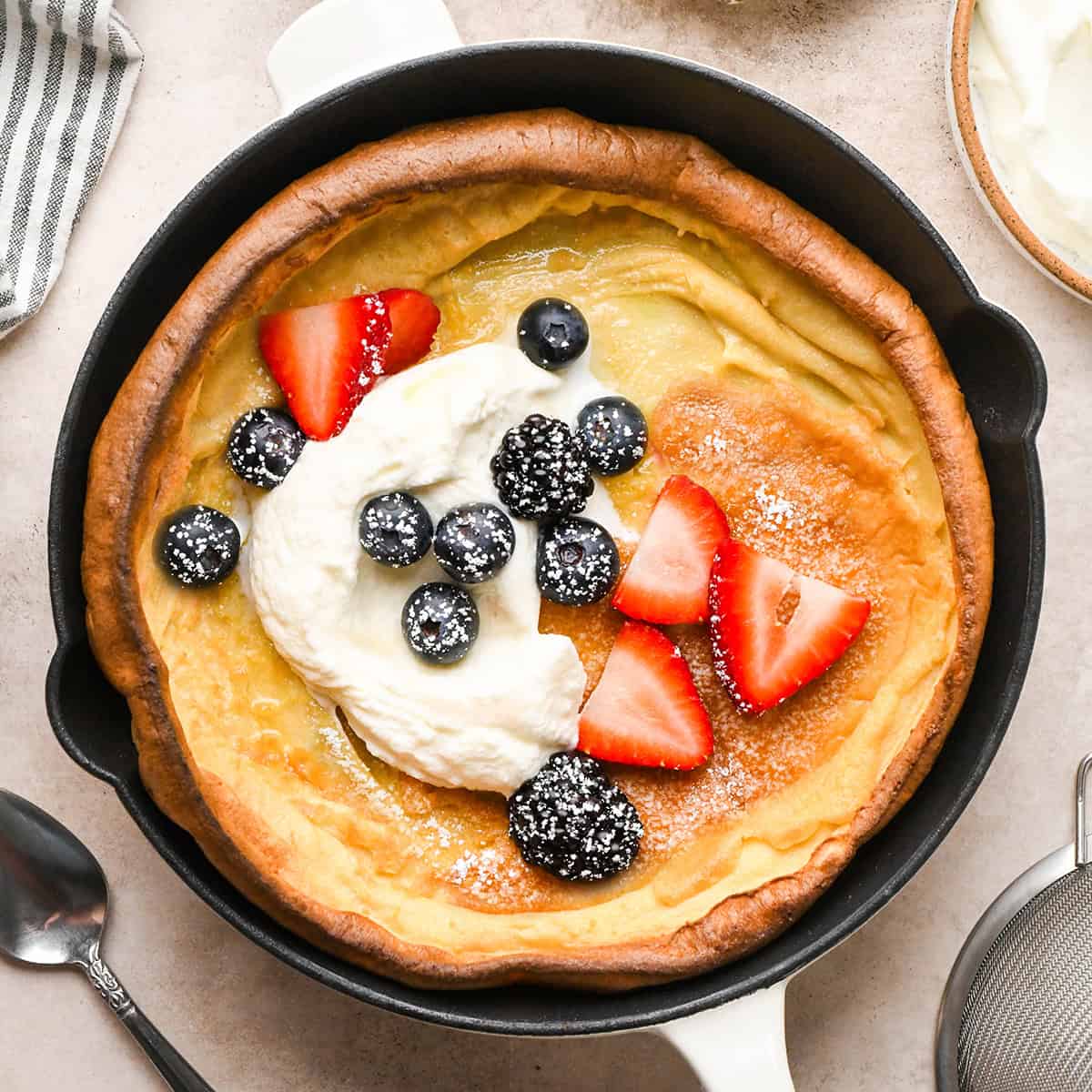 Overhead view of a golden, puffed Dutch baby pancake baked in a cast-iron skillet on a light countertop and topped with strawberries, blueberries, blackberries, whipped cream, and powdered sugar.