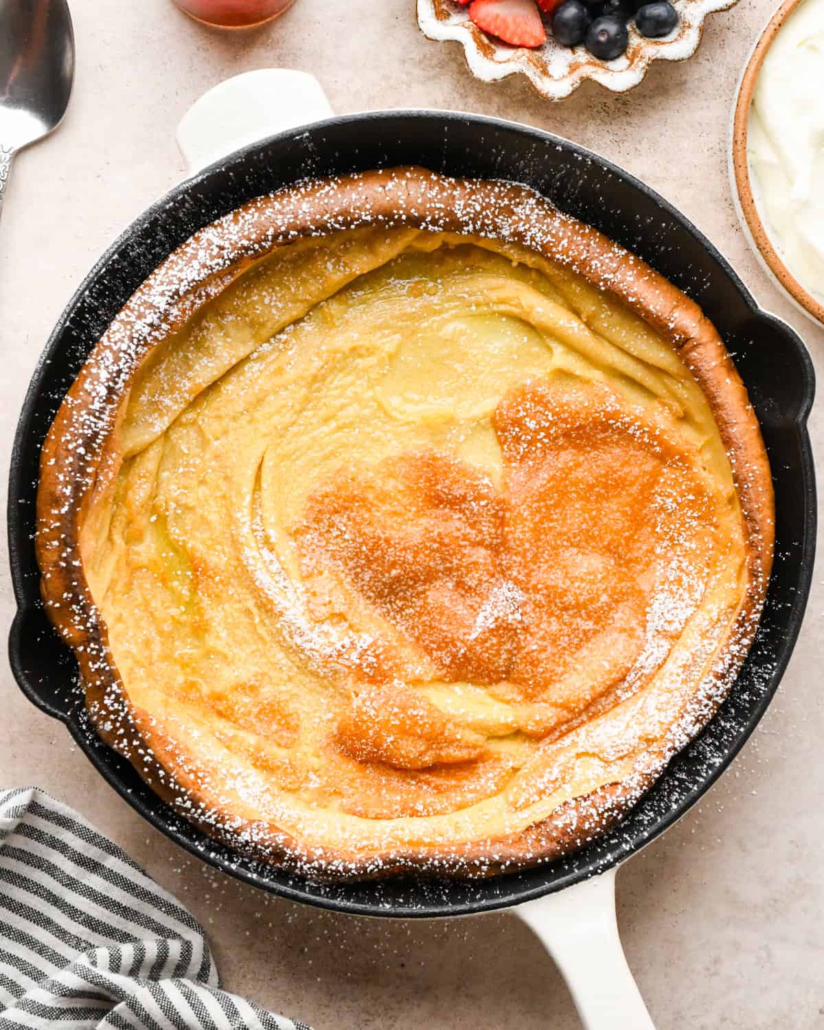 Overhead view of a golden, puffed Dutch baby pancake baked in a cast-iron skillet on a light countertop and dusted with powdered sugar.