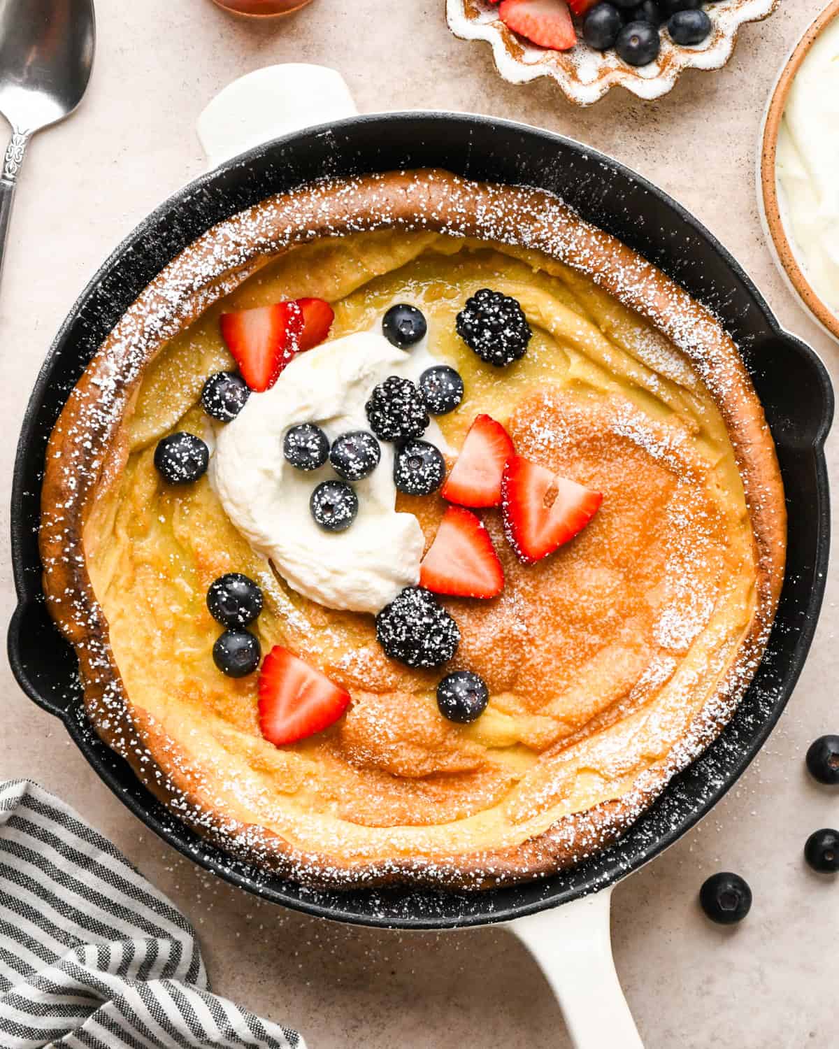 Overhead view of a golden, puffed Dutch baby pancake baked in a cast-iron skillet on a light countertop and topped with strawberries, blueberries, blackberries, whipped cream, and powdered sugar.