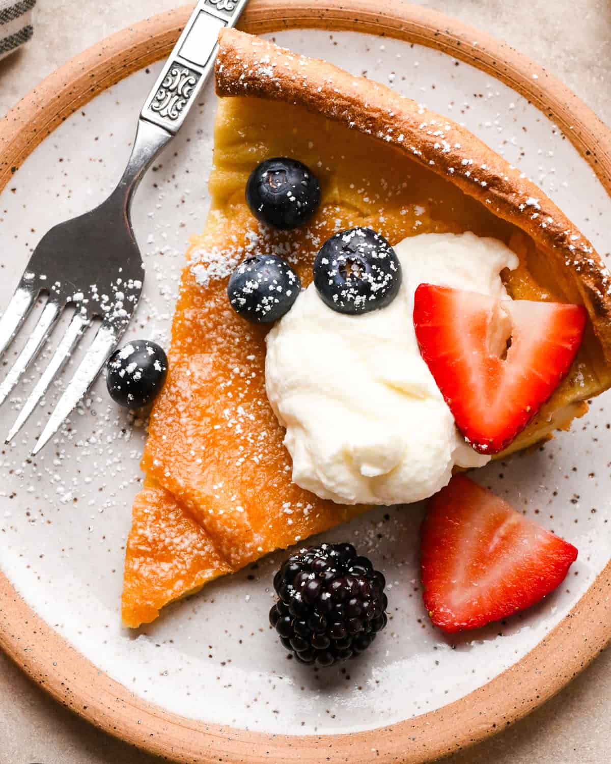 Slice of Dutch baby pancake topped with whipped cream, blueberries, and strawberries, dusted with powdered sugar on a plate with a fork.