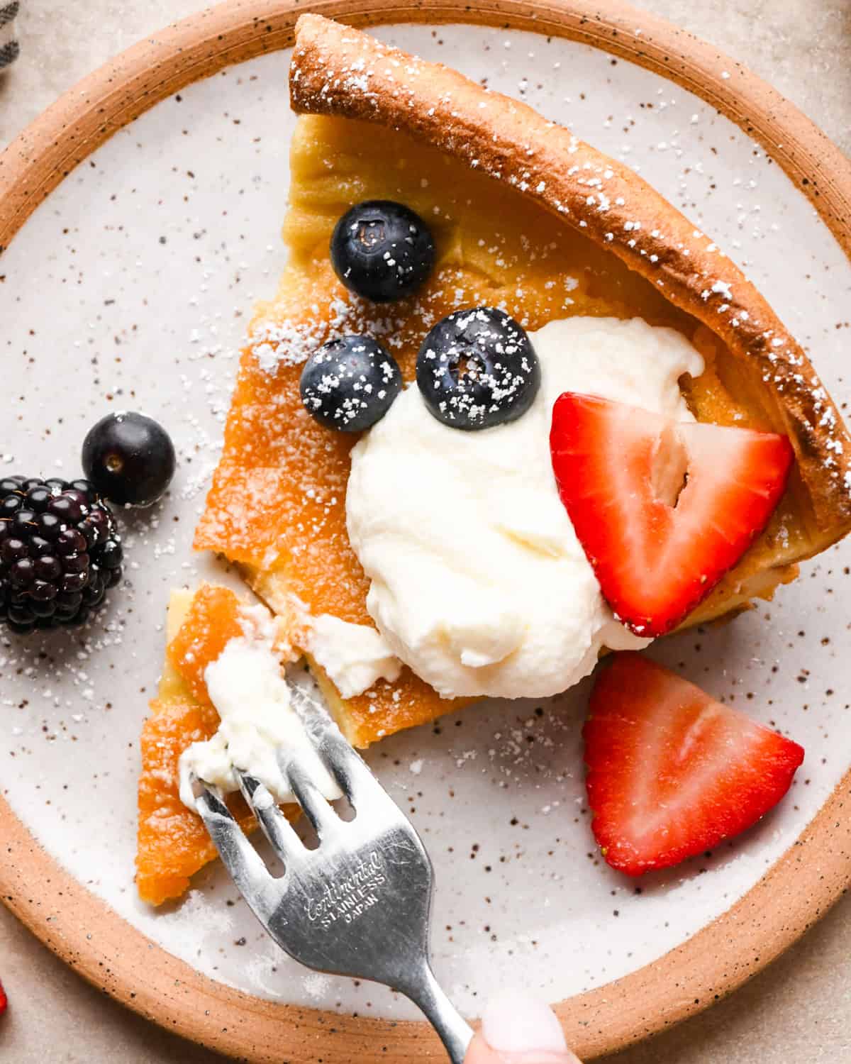 Slice of Dutch baby pancake topped with whipped cream, blueberries, and strawberries, dusted with powdered sugar on a plate with a fork.
