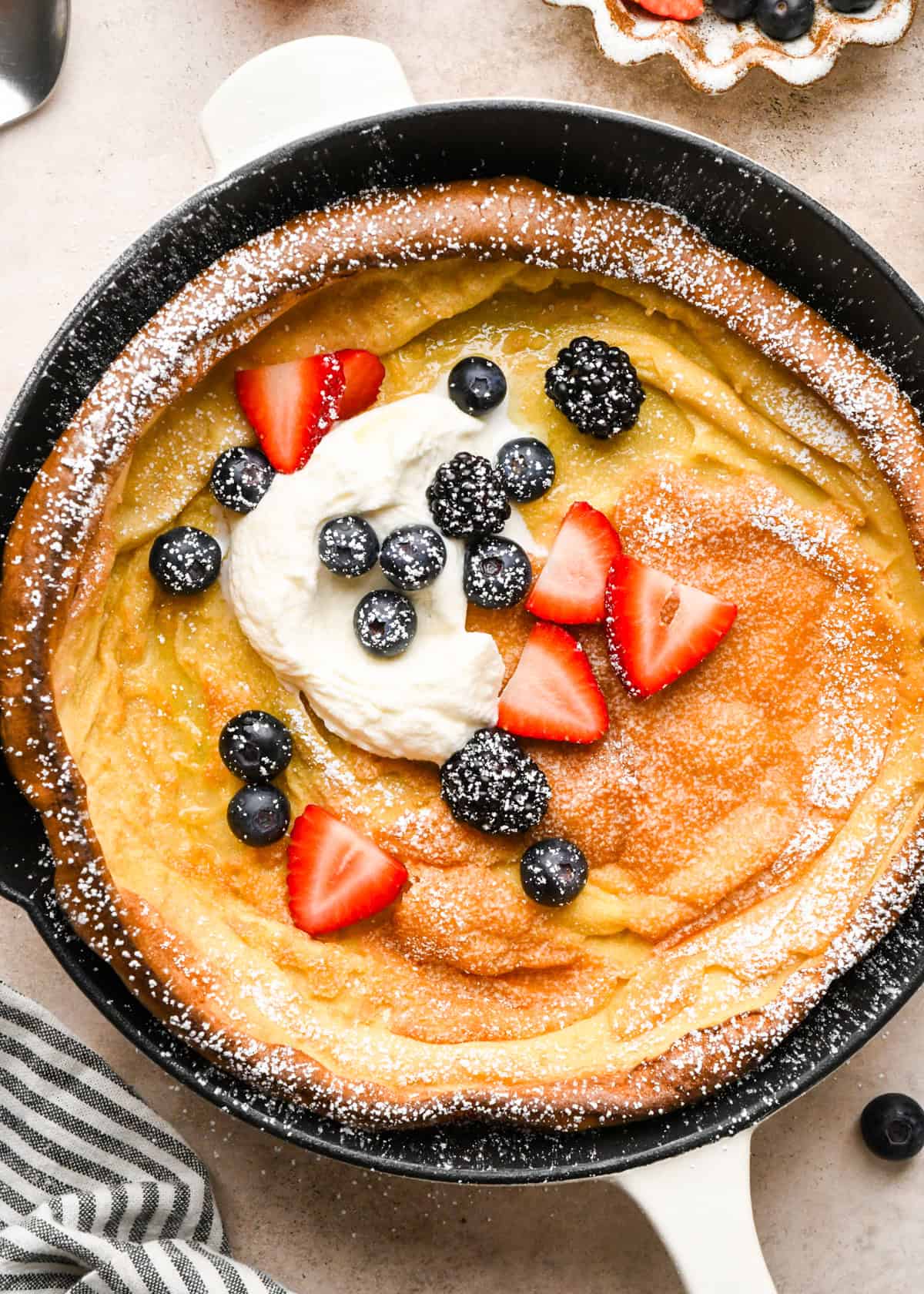Overhead view of a golden, puffed Dutch baby pancake baked in a cast-iron skillet on a light countertop and topped with strawberries, blueberries, blackberries, whipped cream, and powdered sugar.