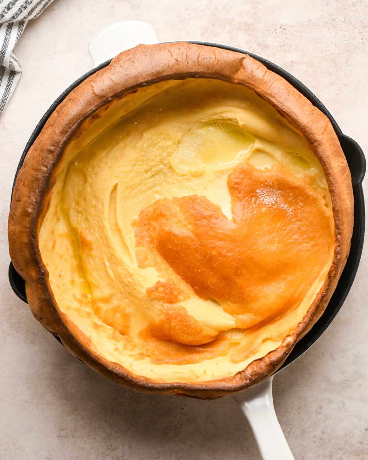Overhead view of a golden, puffed Dutch baby pancake baked in a cast-iron skillet on a light countertop.