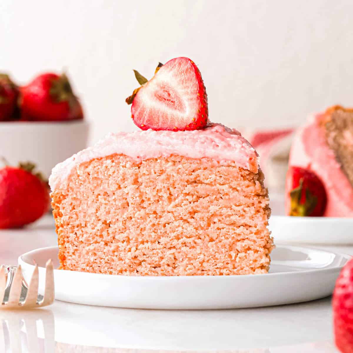 a slice of strawberry cake on a white plate with pink strawberry frosting. There is a half of a strawberry on top of the slice, as well as strawberries on the plate and a bowl of strawberries and another slice of cake in the background.