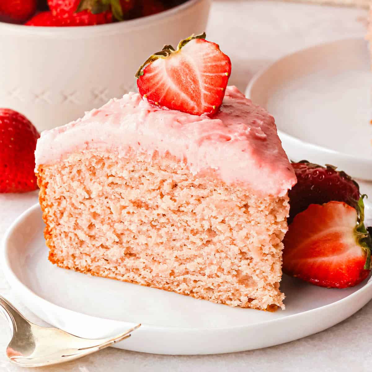 a slice of strawberry cake on a white plate with pink strawberry frosting. There is a half of a strawberry on top of the slice, as well as strawberries on the plate and a bowl of strawberries in the background. There is a fork in the bottom left corner.