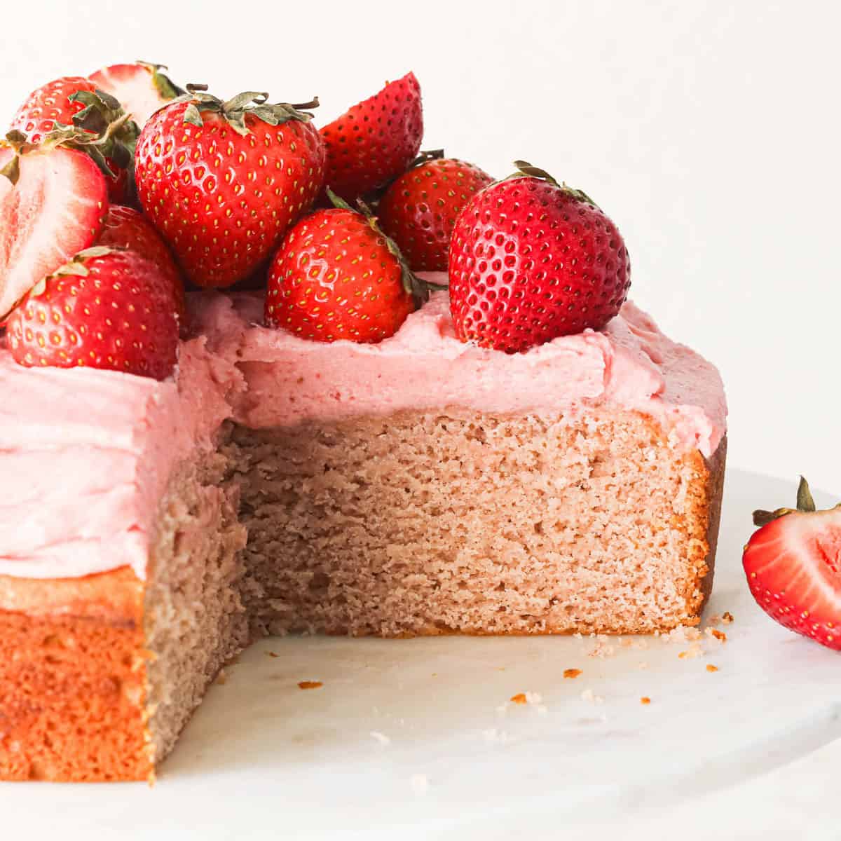 A homemade strawberry cake topped with fresh strawberries and pink strawberry frosting with a slice cut out of it showing the moist interior of the cake. It's on a white cake plate