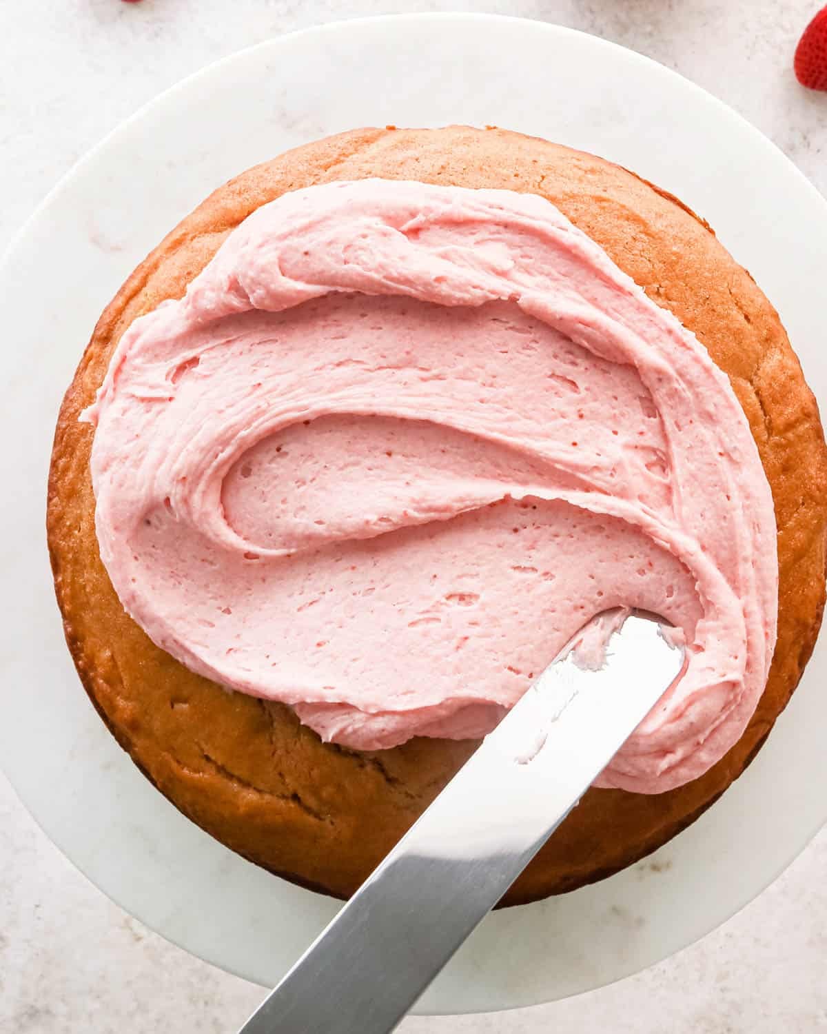 strawberry frosting being spread on top of a round strawberry cake on a white cake plate with a silver spatula.