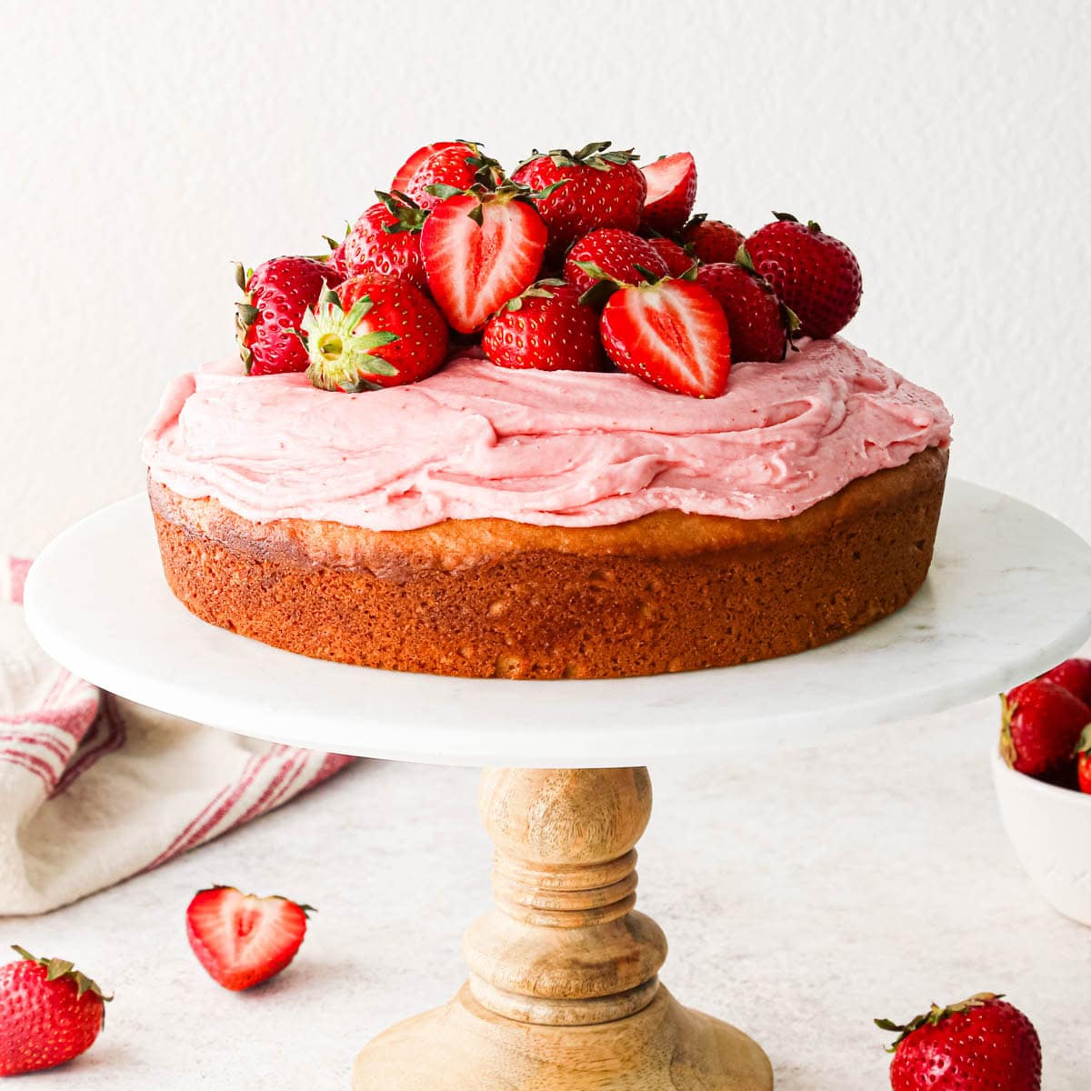 strawberry cake topped with strawberry frosting and a pile of whole and halved strawberries on a marble and wood cake stand surrounded by strawberries.