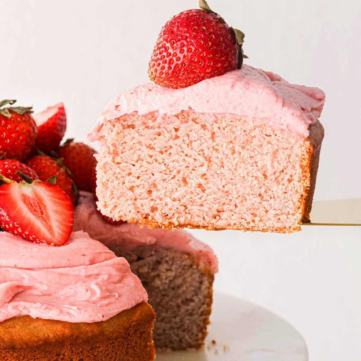 a slice of strawberry cake being lifted off of a cake pan so the moist texture is visible. The cake is topped with pink strawberry frosting and a whole strawberry