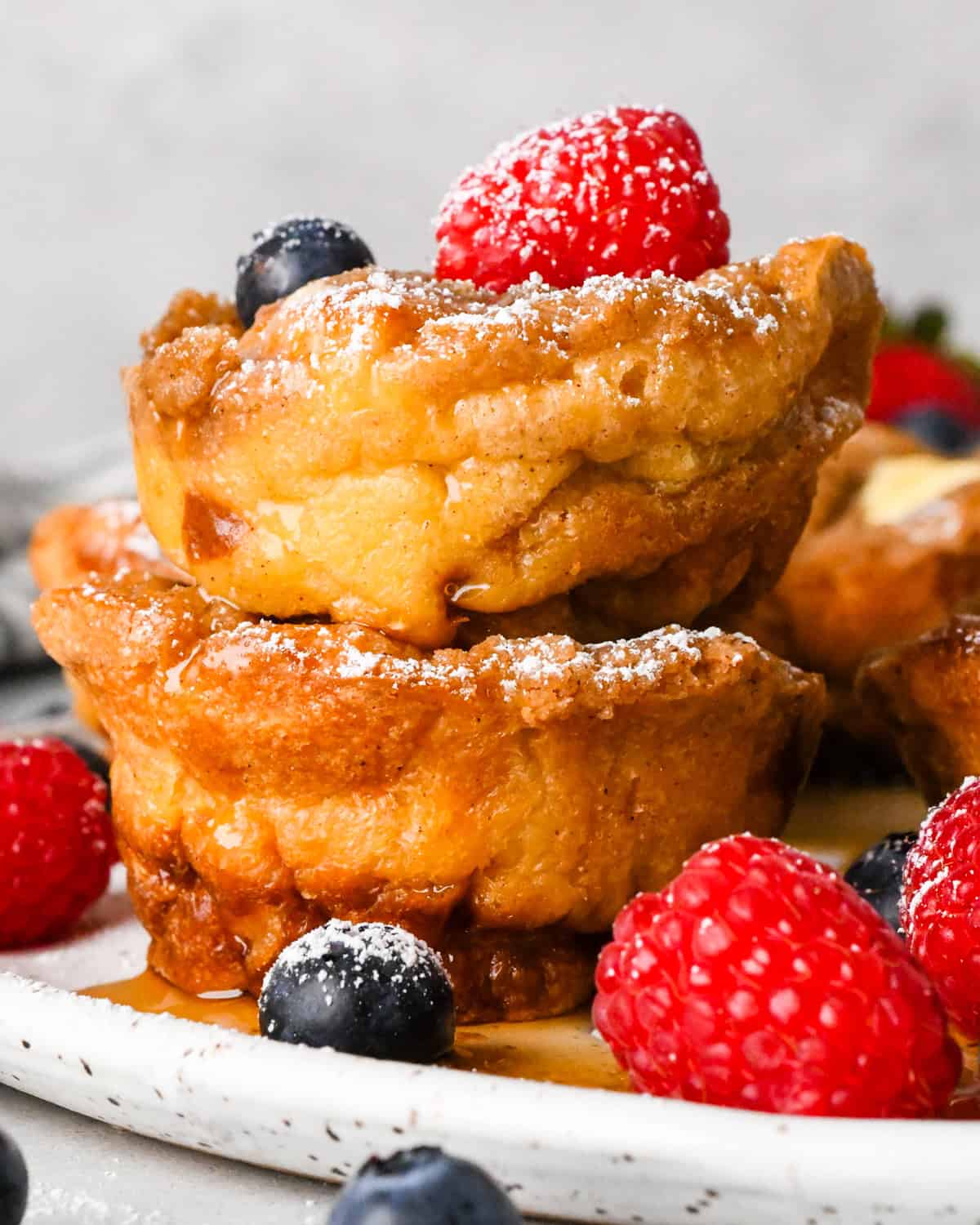 Close-up view of French toast muffins dusted with powdered sugar and topped with melting butter and maple syrup, arranged on a plate with fresh berries.
