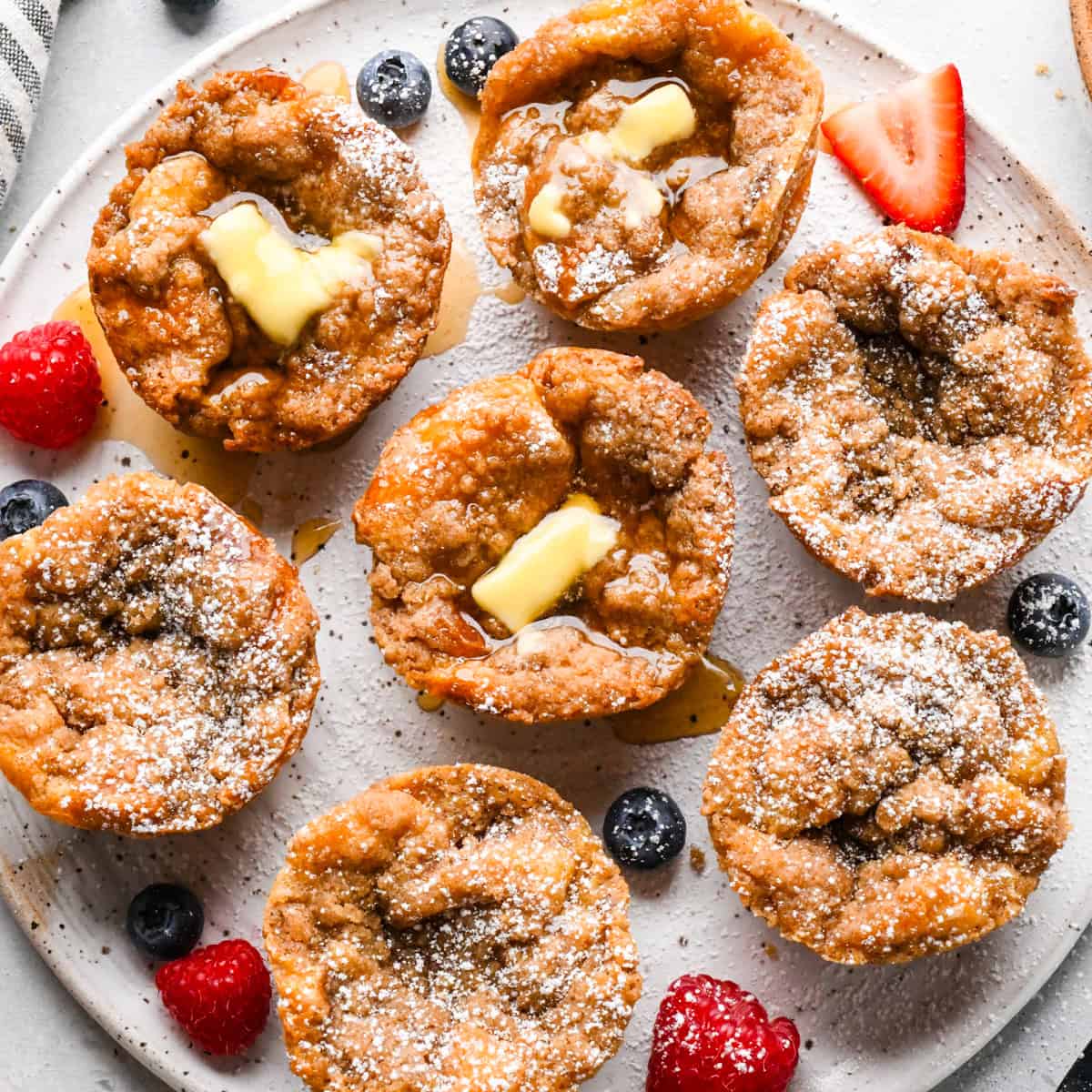 Vue aérienne de muffins de pain perdu saupoudrés de sucre glace et garnis de beurre fondant et de sirop d'érable, disposés sur une assiette avec des baies fraîches.