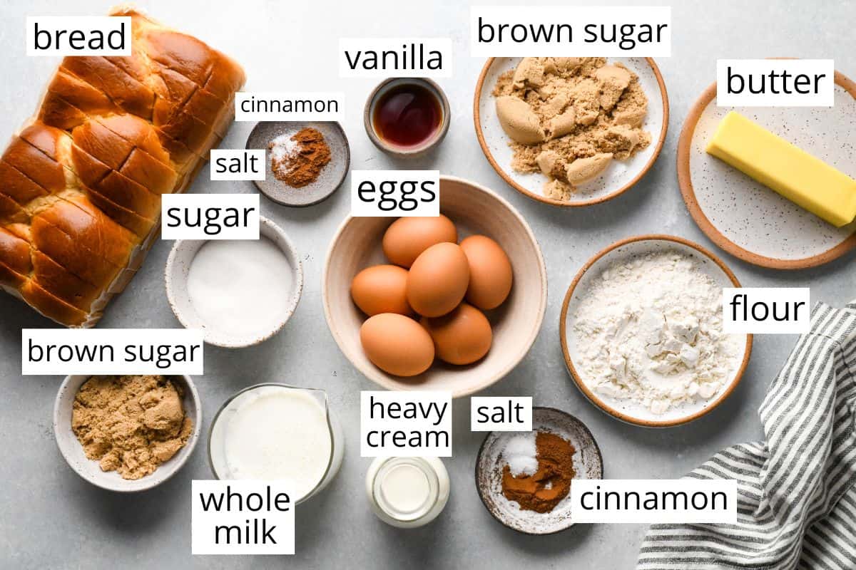 Overhead view of labeled ingredients for French toast muffins arranged on a light surface, including sliced bread, eggs, whole milk, heavy cream, butter, brown sugar, granulated sugar, flour, vanilla, cinnamon, and salt.