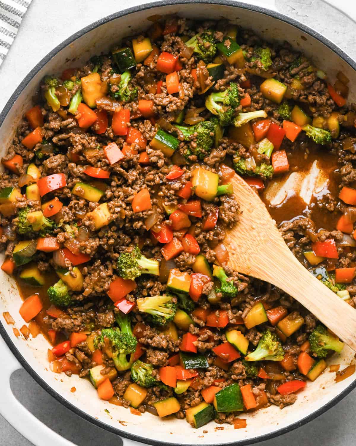 Ground beef stir fry in a skillet with broccoli, red bell peppers, zucchini, and carrots with a wooden spatula in it showing the thick rich sauce