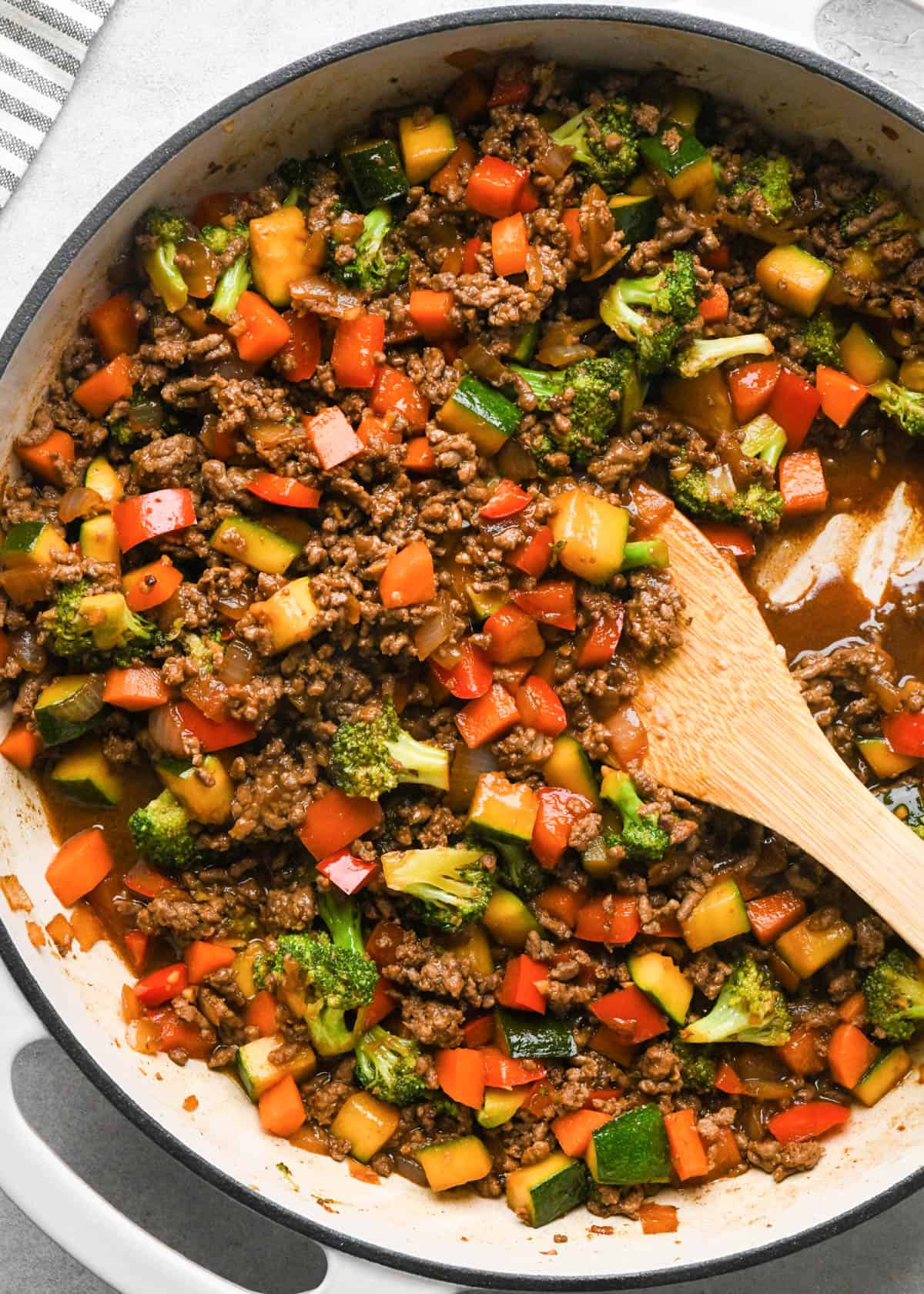 Ground beef stir fry in a skillet with broccoli, red bell peppers, zucchini, and carrots with a wooden spatula in it showing the thick rich sauce
