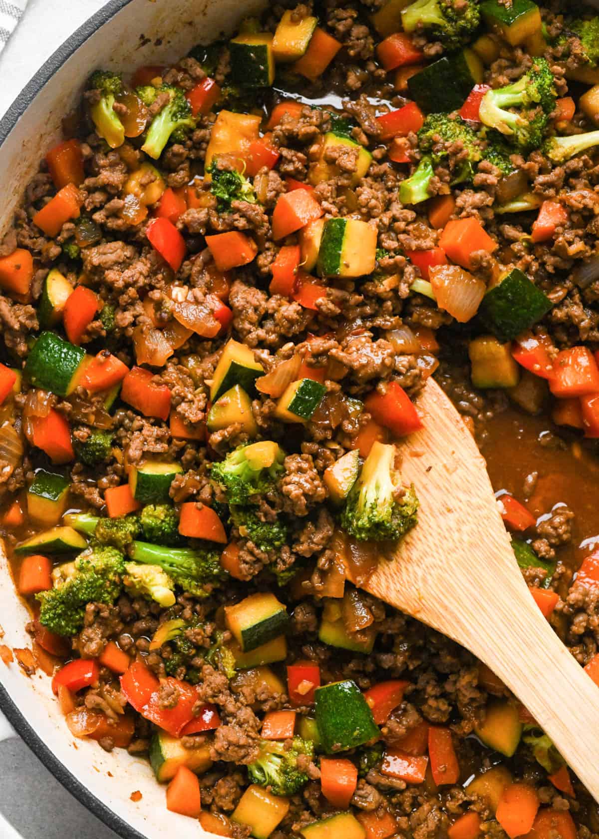 close up photo of Ground beef stir fry in a skillet with broccoli, red bell peppers, zucchini, and carrots with a wooden spatula in it showing the thick rich sauce