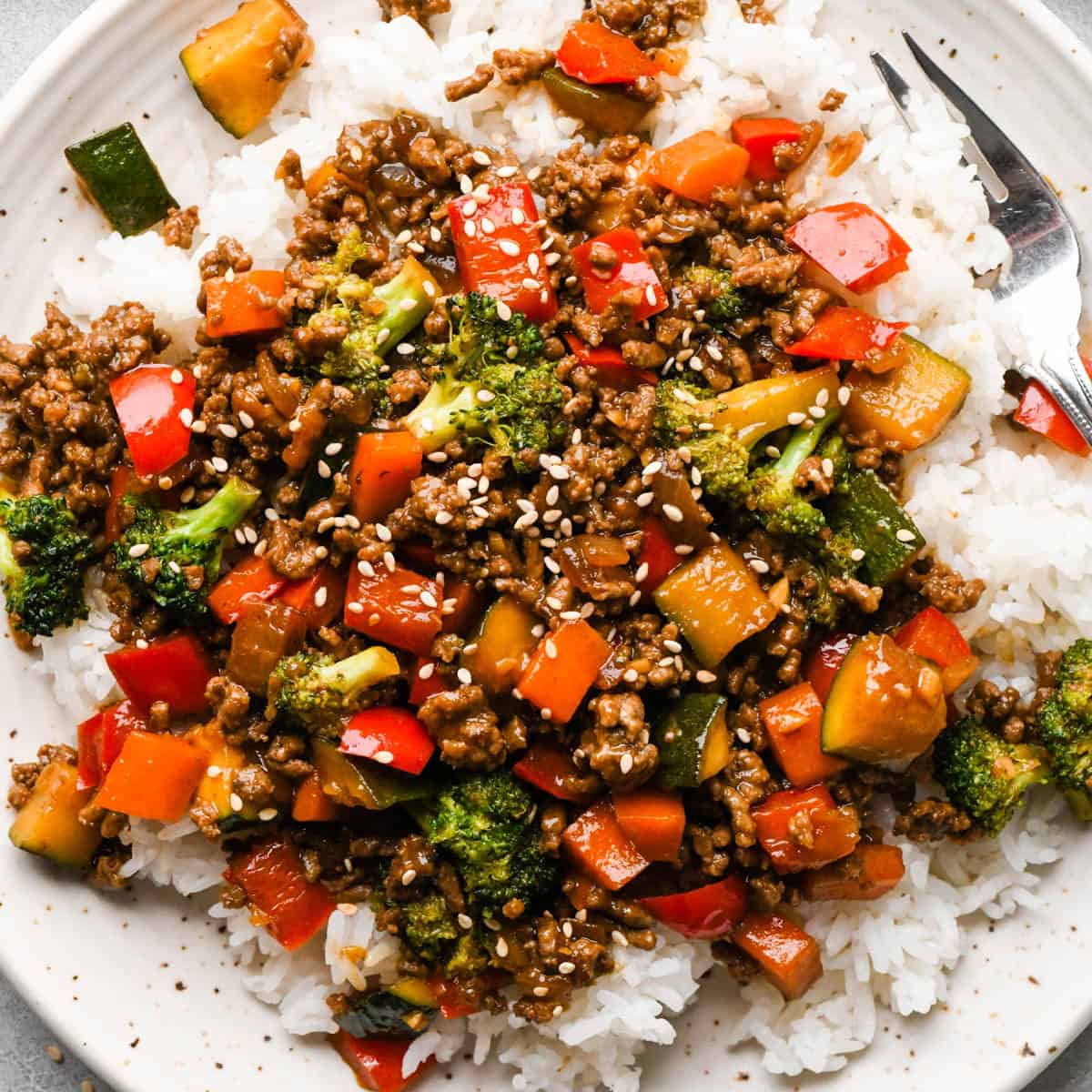 Ground beef stir fry on a white ceramic plate with broccoli, red bell peppers, zucchini, and carrots served over white rice, topped with sesame seeds. there's a fork in the top right corner of the plate