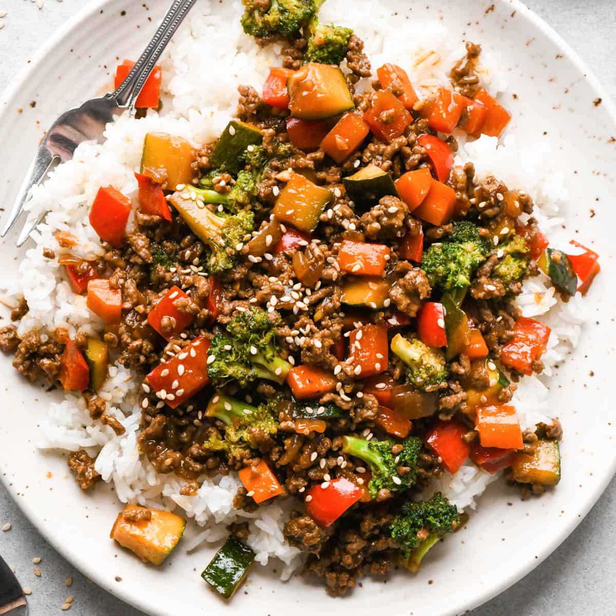 Ground beef stir fry on a white ceramic plate with broccoli, red bell peppers, zucchini, and carrots served over white rice, topped with sesame seeds. there's a fork in the top left corner of the plate