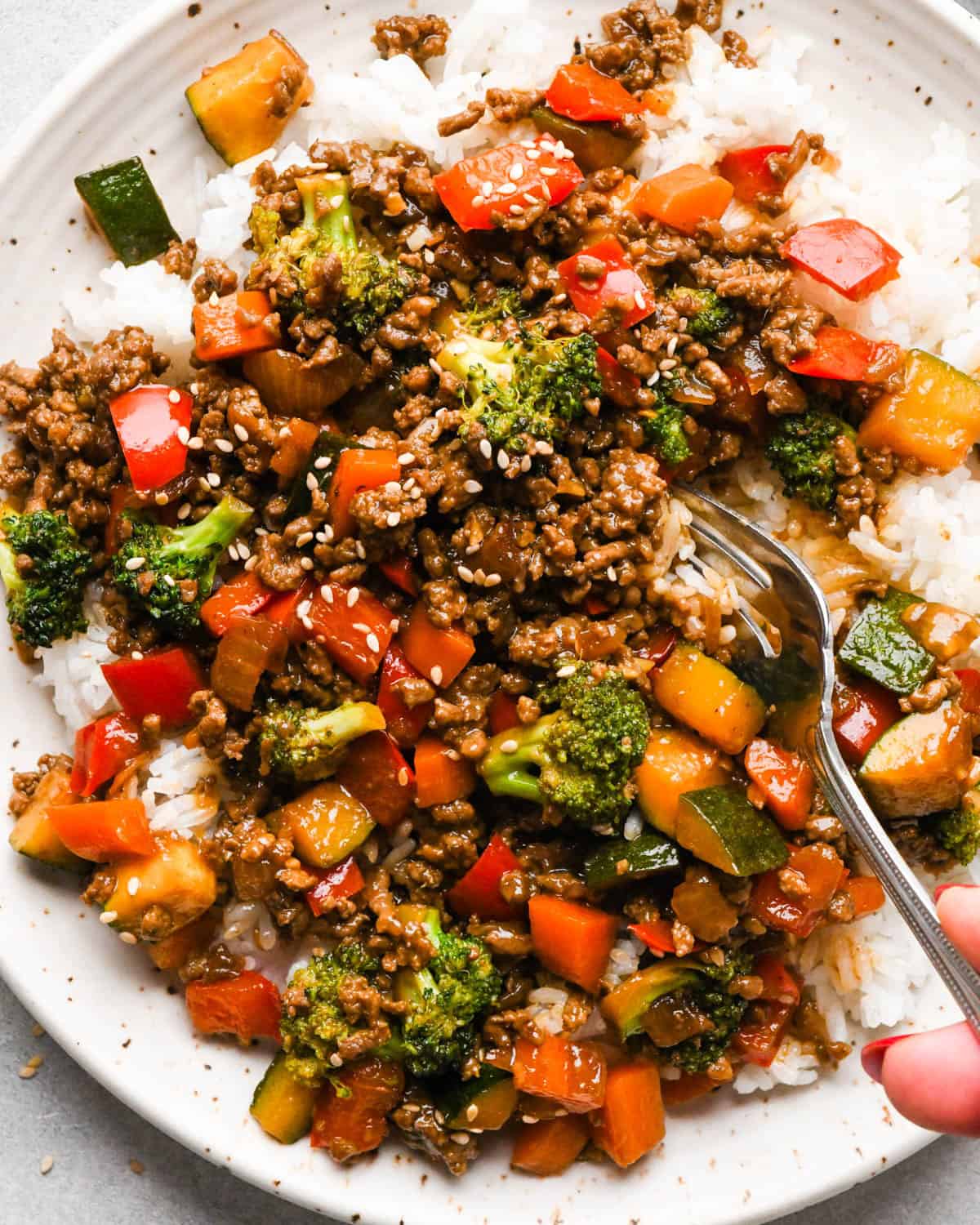 a fork taking a scoop of Ground beef stir fry on a white ceramic plate with broccoli, red bell peppers, zucchini, and carrots served over white rice, topped with sesame seeds