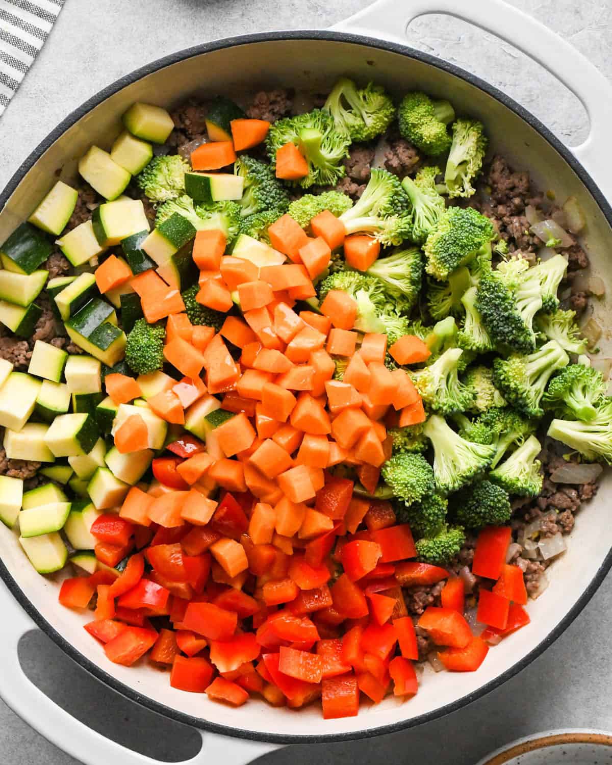 making ground beef stir fry - carrots, zucchini, red bell pepper and broccoli added to the ground beef in a white skillet with a black rim.