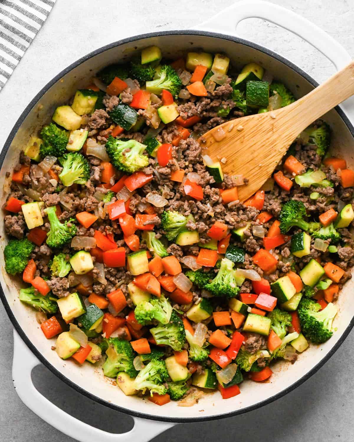 making ground beef stir fry - ground beef and veggies cooked in a white skillet with a black rim and a wooden spatula