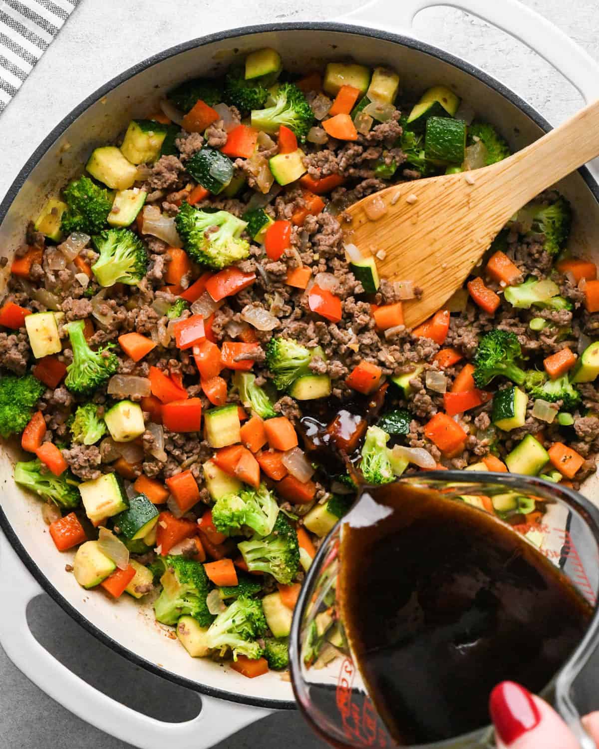 making ground beef stir fry - sauce being poured from a pyrex container over the ground beef and veggies cooked in a white skillet with a black rim and a wooden spatula